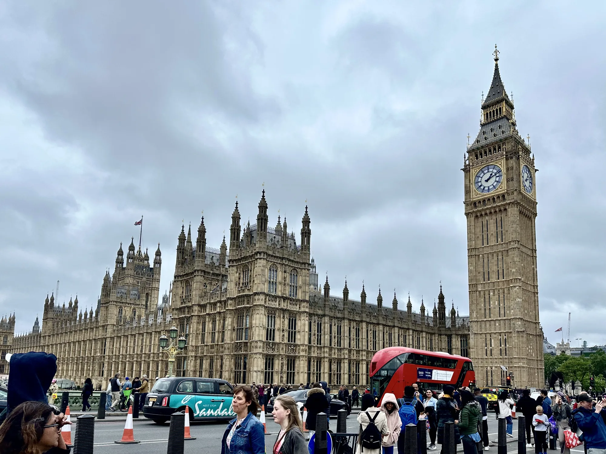 写真はイギリスのロンドンで撮影されたもので、国会議事堂とビッグ・ベン（エリザベス・タワー）が写っています。空はどんよりとした曇り空で、重厚な石造りの建物が印象的です。国会議事堂にはイギリスの国旗が掲げられています。手前には道路があり、人々が行き交っています。ロンドンの象徴的な赤い二階建てバスや、タクシーも走っており、観光地らしい賑わいを見せています。歩道には観光客と思われる人々がおり、写真を撮ったり景色を眺めたりしている様子が伺えます。全体的に落ち着いた色調で、歴史と文化を感じさせる雰囲気です。