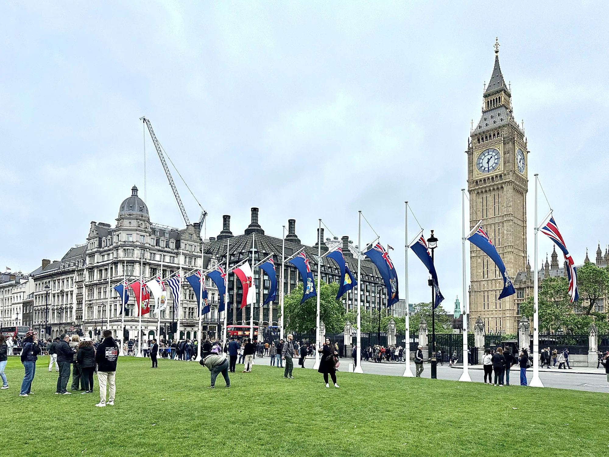 写真は、イギリスのロンドンで撮影された風景です。手前には広々とした緑の芝生が広がっており、人々が散策したり、立ち止まって談笑したりしています。芝生の奥には、様々な国の旗がポールに掲げられており、風になびいています。  背景には、ロンドンの象徴的な建造物であるビッグ・ベン（エリザベス・タワー）がそびえ立っています。また、その隣には歴史を感じさせる建物が立ち並んでおり、重厚な雰囲気を醸し出しています。さらに、建設中のクレーンが写っており、ロンドンの街が常に変化し続けていることを示唆しています。  全体的に曇り空で、落ち着いた雰囲気の写真です。観光客や地元の人々が集まり、ロンドンの歴史と現代が融合した風景を楽しんでいる様子が伝わってきます。