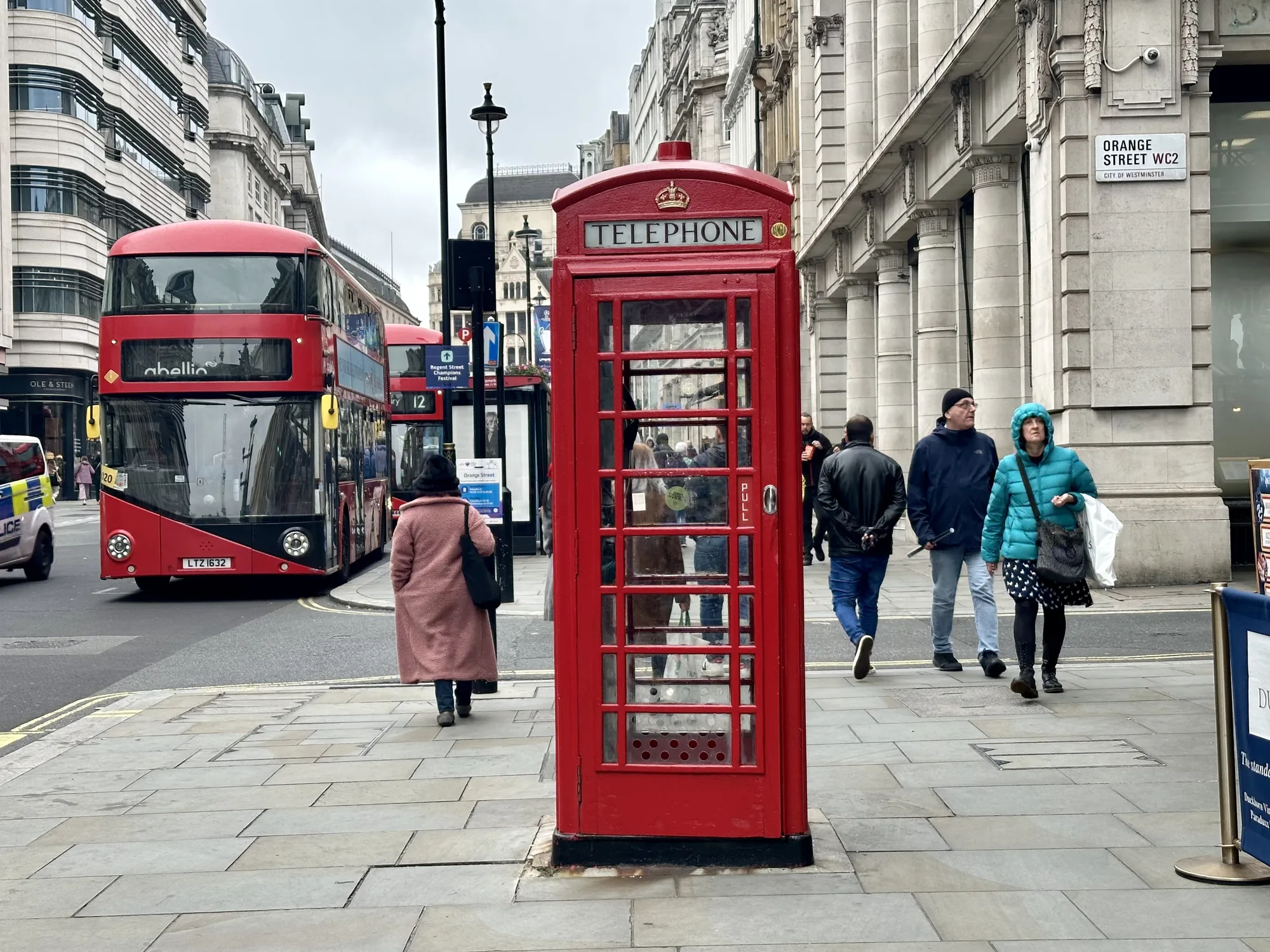 写真はイギリスのロンドンで撮影された街並みを捉えています。中央には、イギリスの象徴とも言える赤い電話ボックスが置かれています。電話ボックスの背後には、他の通行人がぼんやりと映り込んでいます。  画面左側には赤い二階建てバスが走行しており、その隣には警察車両が停車しています。道路の反対側には、上品な石造りの建物が立ち並んでいます。写真全体は曇り空の下で撮影されており、落ち着いた雰囲気を醸し出しています。歩道には人々が歩いており、日常の風景が切り取られています。右側には「ORANGE STREET WC2」と書かれた道路標識が見えます。全体的に、ロンドンの典型的な街の様子を捉えた写真と言えるでしょう。
