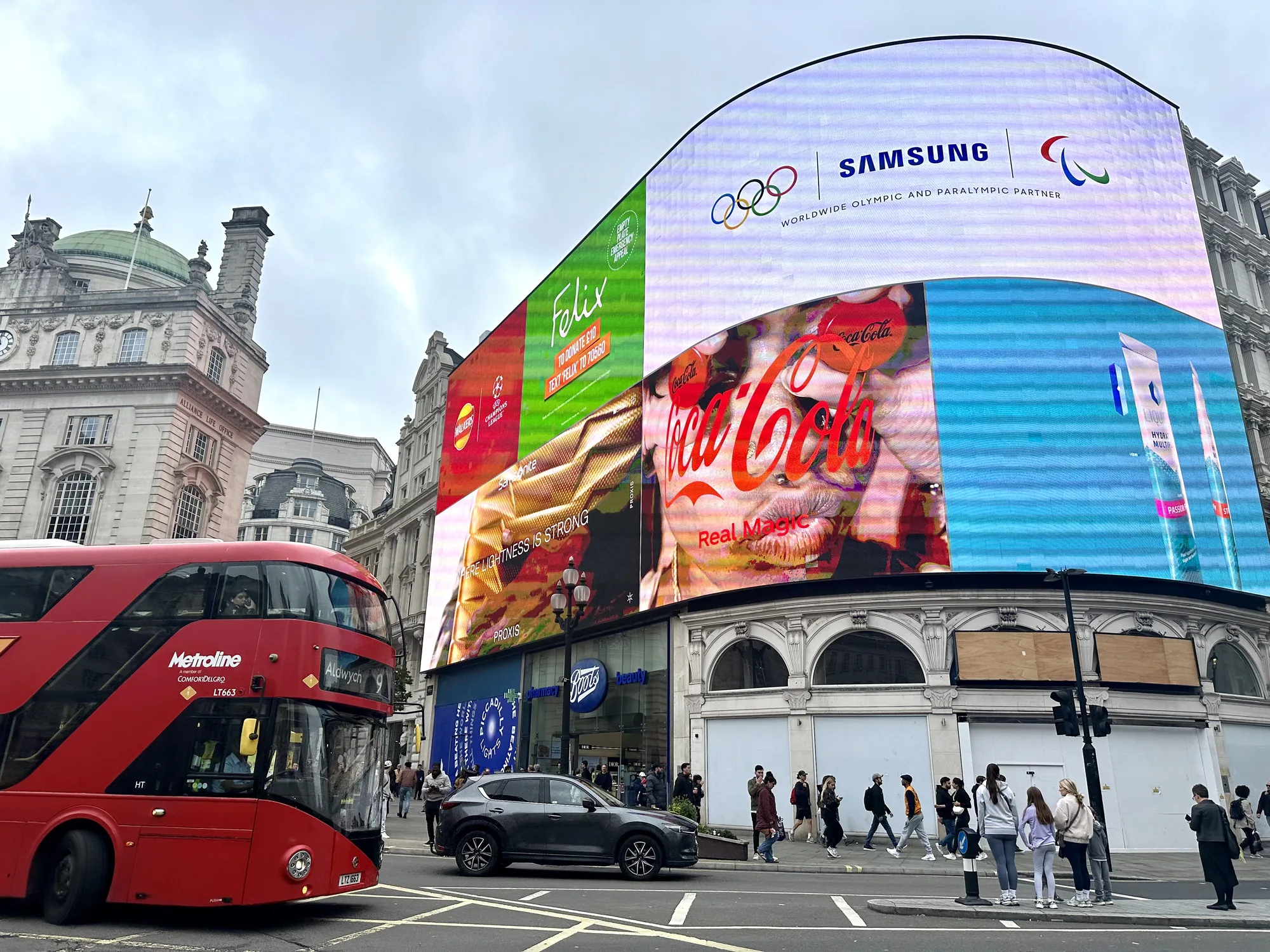 写真は、ロンドンの有名な観光名所であるピカデリーサーカスを捉えています。空は曇りで、落ち着いた雰囲気です。正面には赤い2階建てバスが走り、ロンドンの象徴的な風景を演出しています。背景には、巨大な広告スクリーンがそびえ立ち、コカ・コーラ、サムスン、フェリックス、サムソナイト、WALKERS、PROXISなど、様々な企業の広告が表示されています。サムスンの広告はオリンピックとパラリンピックのパートナーシップを示しています。歩道には人々が行き交い、様々な服装をしています。Bootsの店舗も確認できます。建物の壁は改装中のようです。