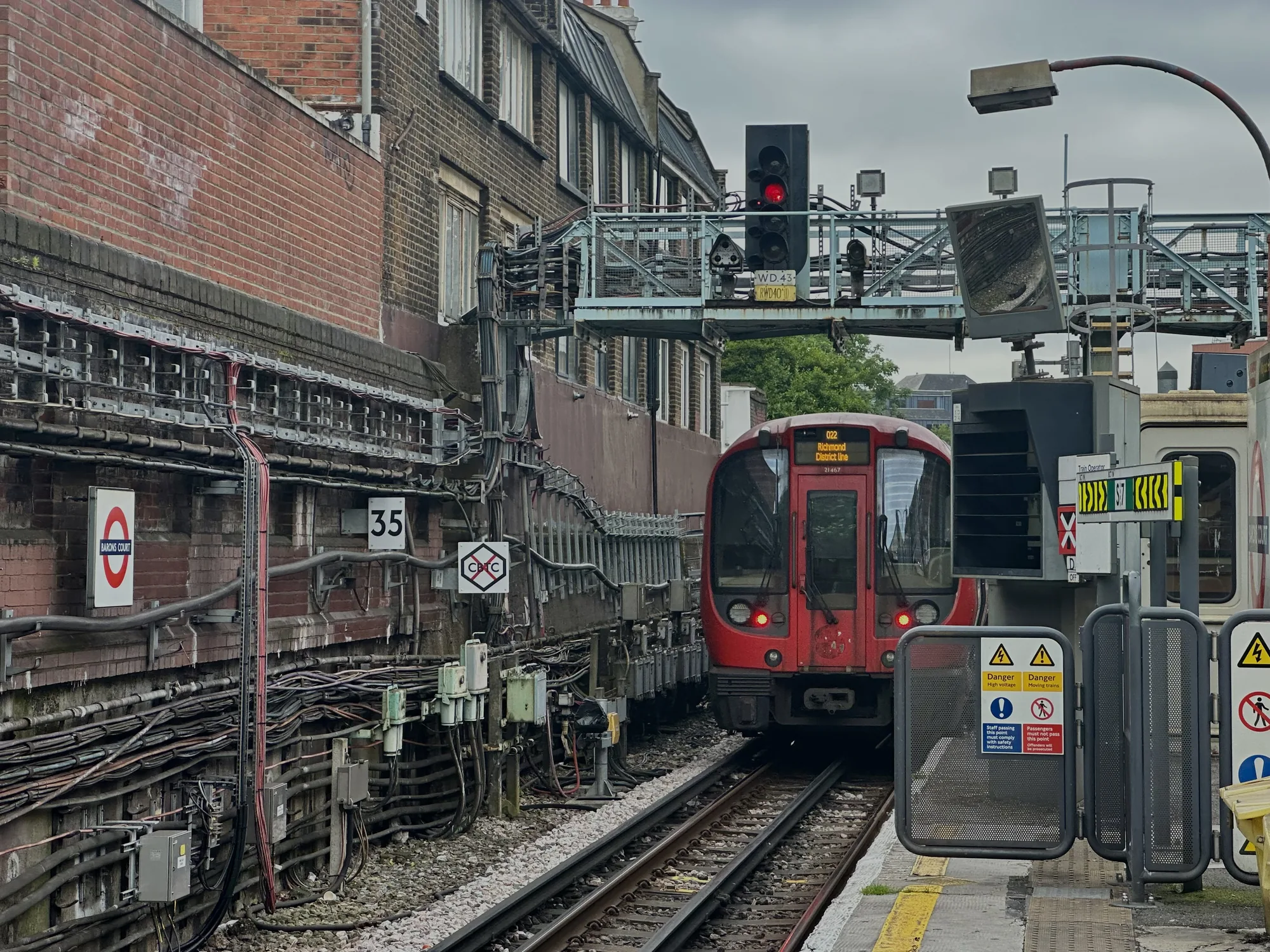 写真はイギリス、ロンドンの地下鉄駅の様子を捉えています。赤い車体の地下鉄車両がホームに停車しており、行先表示には「Richmond District Line」と書かれています。線路の奥には赤信号が点灯しており、列車が停止している理由を示唆しています。駅の壁面はレンガ造りで、無数のケーブルや配管が張り巡らされており、都市のインフラの複雑さを感じさせます。また、「Barons Court」という駅名を示す円形のロゴや、番号「35」が壁に掲げられています。ホームには安全柵が設置され、「Danger High voltage」「Danger Moving trains」などの注意喚起の表示が見られます。全体的に曇り空で、やや暗く落ち着いた雰囲気です。