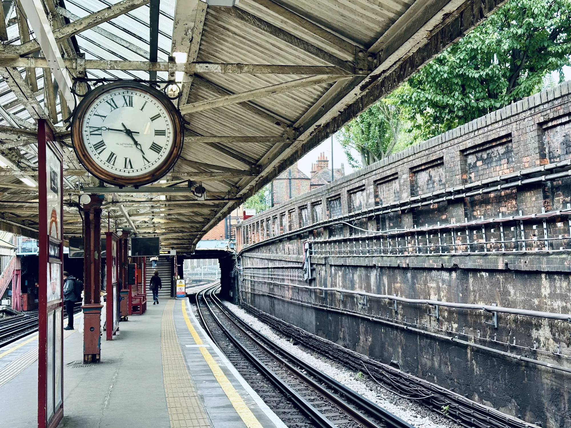 写真は、イギリスのロンドンの鉄道駅のプラットフォームを捉えたものです。天井には古びた白い鉄骨の構造があり、ガラスがはめ込まれています。その下には、文字盤がローマ数字の大きなアナログ時計が吊るされています。プラットフォームはグレーのコンクリートで、黄色い線が引かれています。柱は赤茶色に塗られ、広告などが掲示されています。数人の人がプラットフォームを歩いています。線路はカーブを描き、トンネルへと続いています。線路脇には、レンガの壁がそびえ立ち、壁には無数のケーブルが這っています。壁の上には緑の木々が見え、都会の中にも自然が感じられます。全体的に、ややくすんだ色調で、歴史を感じさせる雰囲気が漂っています。