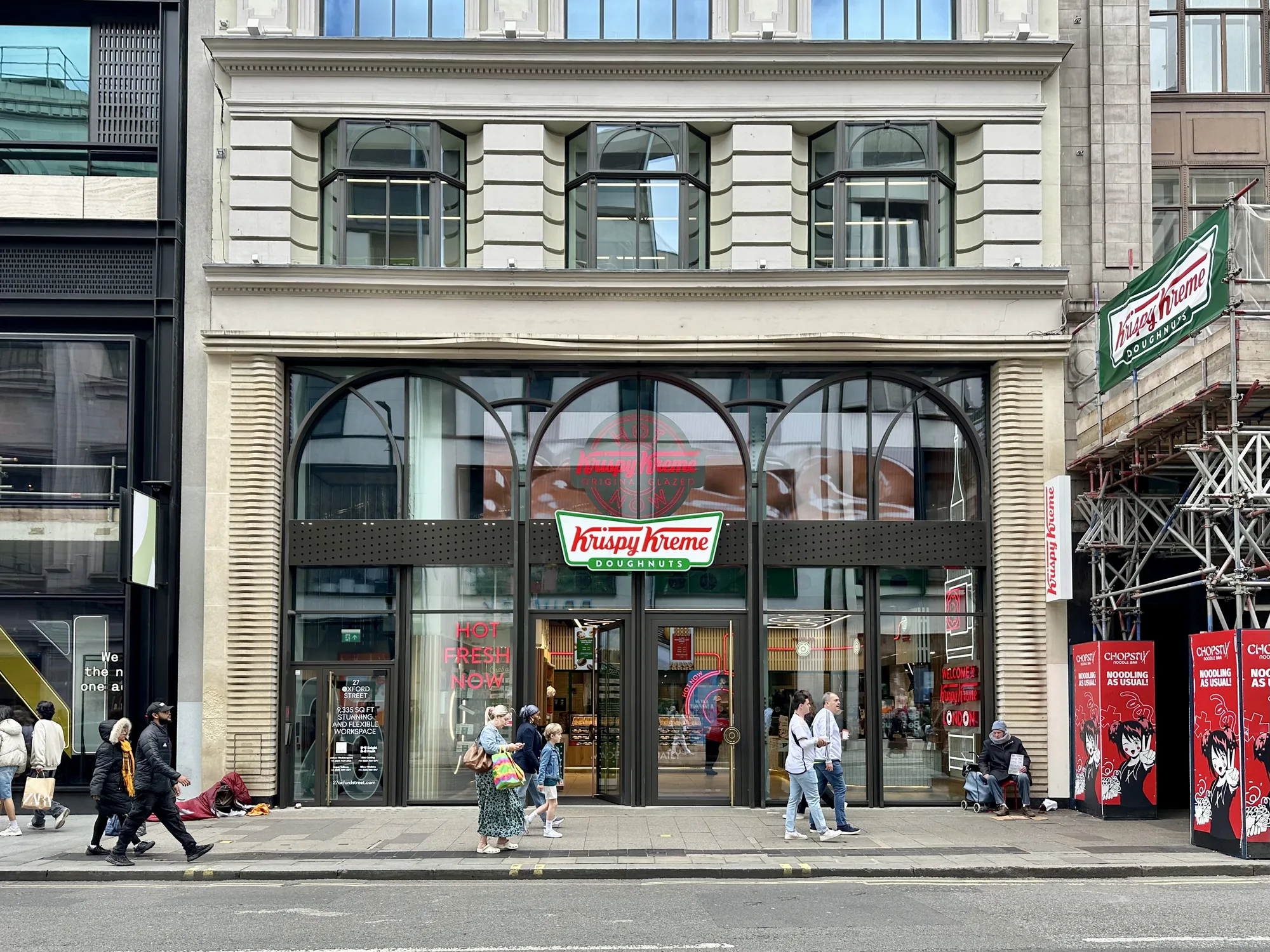 この写真はイギリスのロンドンの街並みを捉えたものです。中心には「Krispy Kreme DOUGHNUTS」の店舗があり、大きなアーチ型の窓が特徴的です。店舗の入り口には「HOT FRESH NOW」という赤いネオンサインが見えます。歩道には数人の人が歩いており、中には店に入る人もいます。また、店舗の隣には工事現場のような足場が組まれており、「Krispy Kreme」の看板が取り付けられています。歩道には物乞いをしている人もいます。全体的に都会の賑やかな様子が伝わってきます。