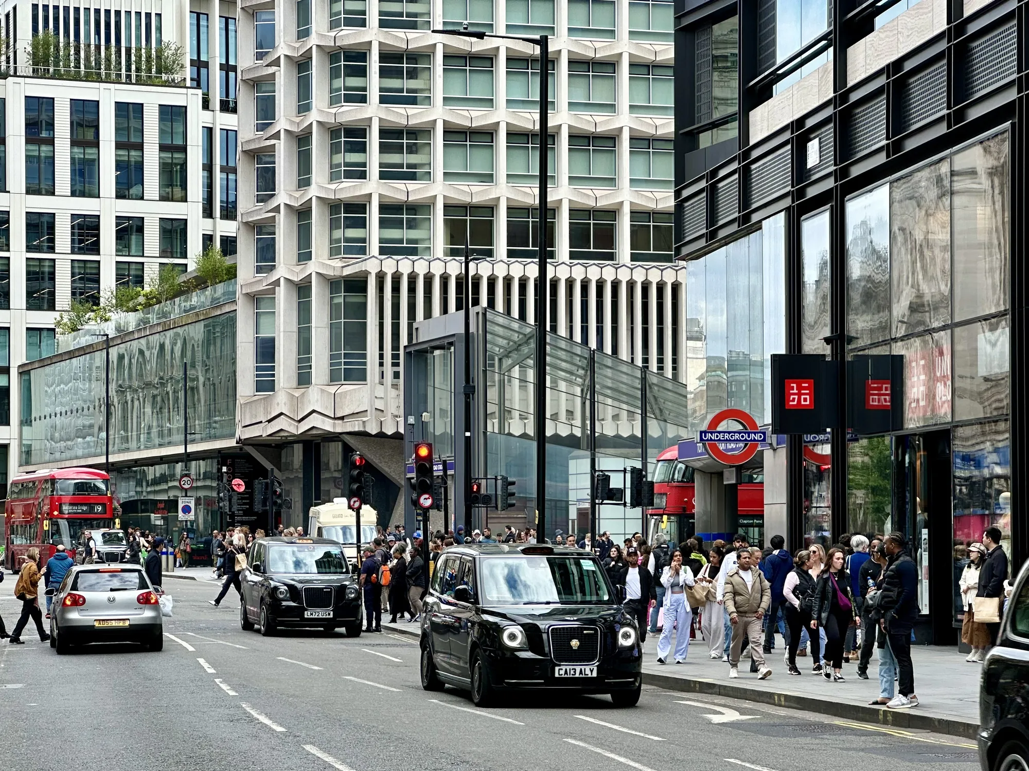 写真は、イギリスのロンドンで撮影された都市の風景を捉えています。通りには多くの人が行き交い、活気にあふれています。道路にはロンドンの象徴的なブラックキャブが複数台走っており、赤い二階建てバスも確認できます。歩道には人々が密集しており、ショッピングや観光を楽しんでいる様子が伺えます。  背景には、近代的なオフィスビルや、特徴的なデザインの建物が立ち並んでいます。建物の窓には周囲の風景が反射しており、都市の複雑さを表現しています。地下鉄の駅の標識も見え、ロンドンの公共交通機関の充実ぶりを物語っています。全体的に曇り空で、落ち着いた色調の写真です。