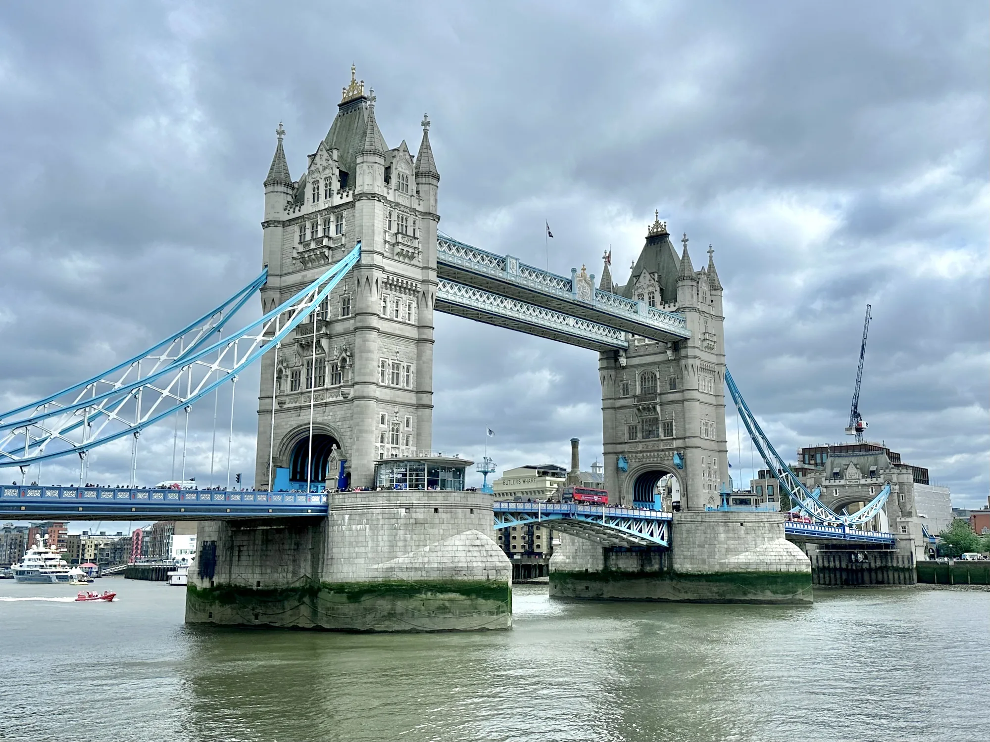 写真には、曇り空の下にそびえ立つロンドンのタワーブリッジが写っています。橋は重厚な石造りの塔を持ち、青色の吊りケーブルが印象的です。橋の上には人々や赤い2階建てバスが往来しており、活気を感じさせます。川面は穏やかで、橋脚には緑色の苔のようなものが付着しています。背景には他の建物やクレーンが見え、都市景観の一部を形成しています。全体的に、曇り空が少し重い雰囲気を醸し出していますが、タワーブリッジの壮大さがそれを打ち消し、ロンドンのランドマークとしての存在感を強く示しています。