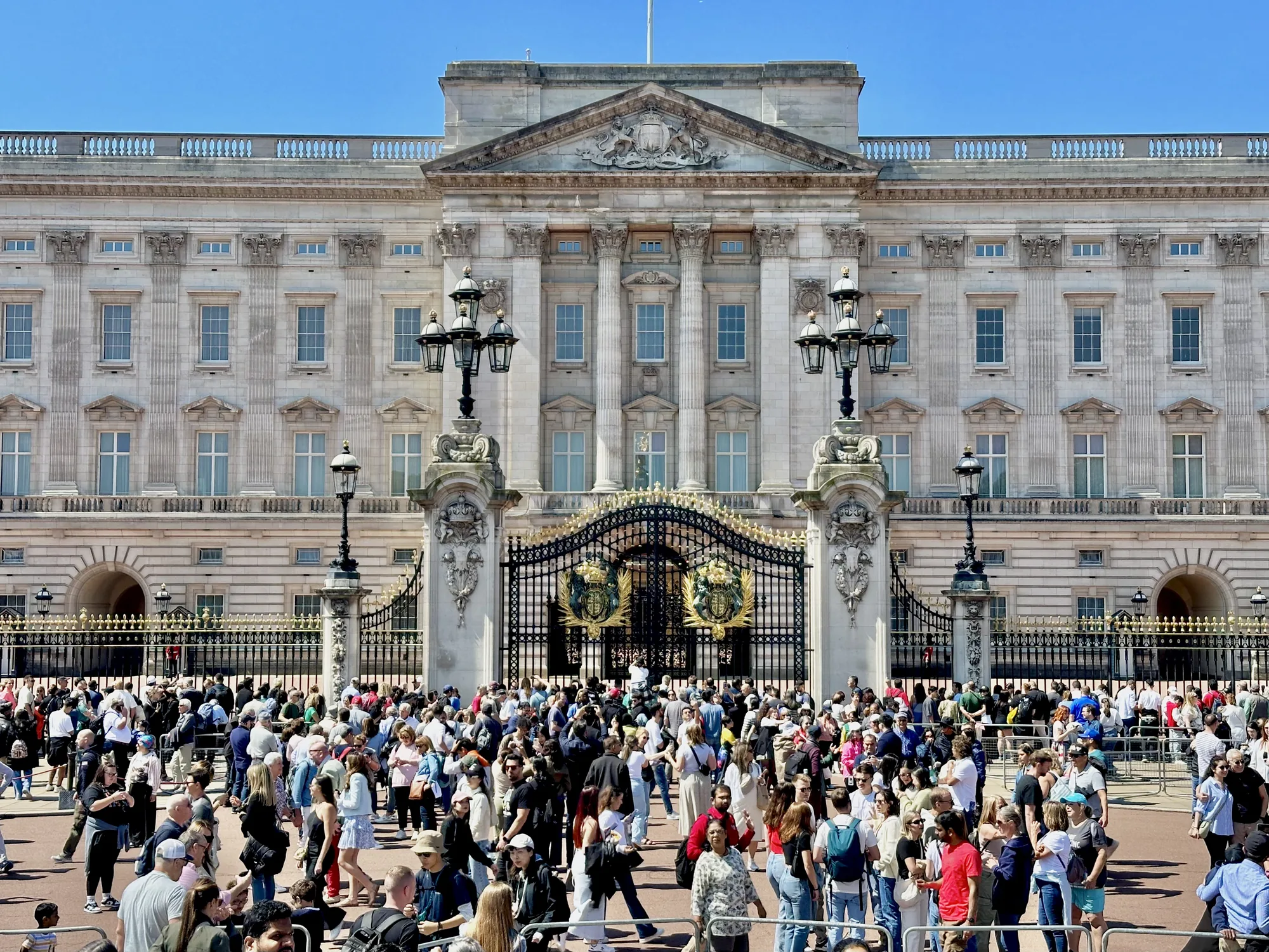 写真は、イギリスのロンドンにあるバッキンガム宮殿を正面から捉えたものです。青空の下、白亜の宮殿が荘厳にそびえ立っています。宮殿の正面には、黒と金色の装飾が施されたゲートがあり、その前には大勢の観光客が集まっています。観光客は様々な服装をしており、写真を撮ったり、景色を楽しんだりしている様子が伺えます。写真全体は明るく、観光地としての活気と賑わいを伝えています。宮殿の建築様式や装飾の細部まで鮮明に写されており、その歴史と風格を感じさせます。