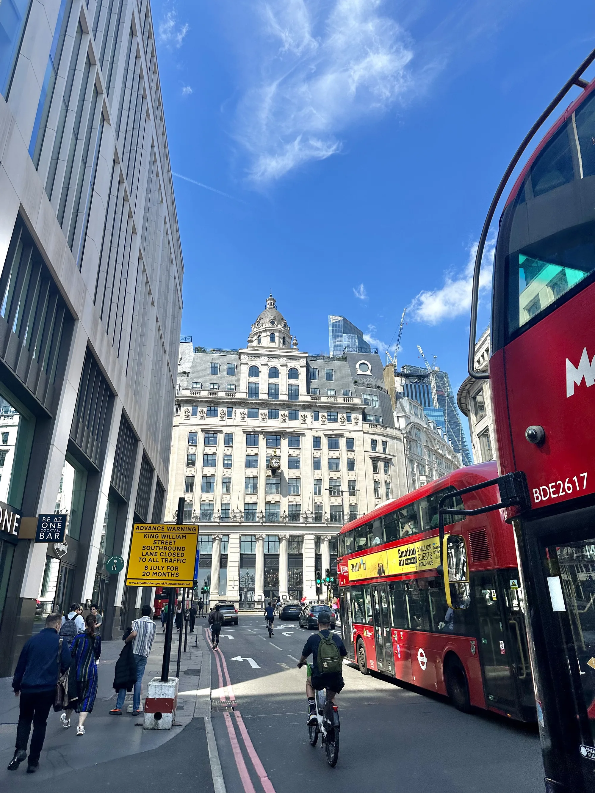 写真はイギリスのロンドンで撮影されたもので、明るい日中の街並みを捉えています。画面右側には、ロンドン名物の赤い二階建てバスの一部が大きく写っており、「M」と「BDE2617」の文字が見えます。その奥には、同じバスの全体像が見え、黄色い広告が描かれています。「Emotion」という文字や、300万人以上のゲストがいるという情報も読み取れます。  道路には自転車に乗った人が一人写っており、車も数台走っています。歩道には数人の歩行者がおり、それぞれ異なる方向に歩いています。  道路脇には黄色い標識があり、「ADVANCE WARNING」と書かれています。「KING WILLIAM STREET SOUTHBOUND LANE CLOSED TO ALL TRAFFIC 8 JULY FOR 20 MONTHS」と書かれており、道路工事による交通規制の情報が伝えられています。  背景には、近代的な高いオフィスビルと、歴史を感じさせる石造りの建物が混在しています。石造りの建物には時計が設置されており、上部にはドーム型の屋根が見えます。空は晴れており、白い雲が浮かんでいます。全体的に、ロンドンの活気ある街の様子が伝わってくる写真です。
