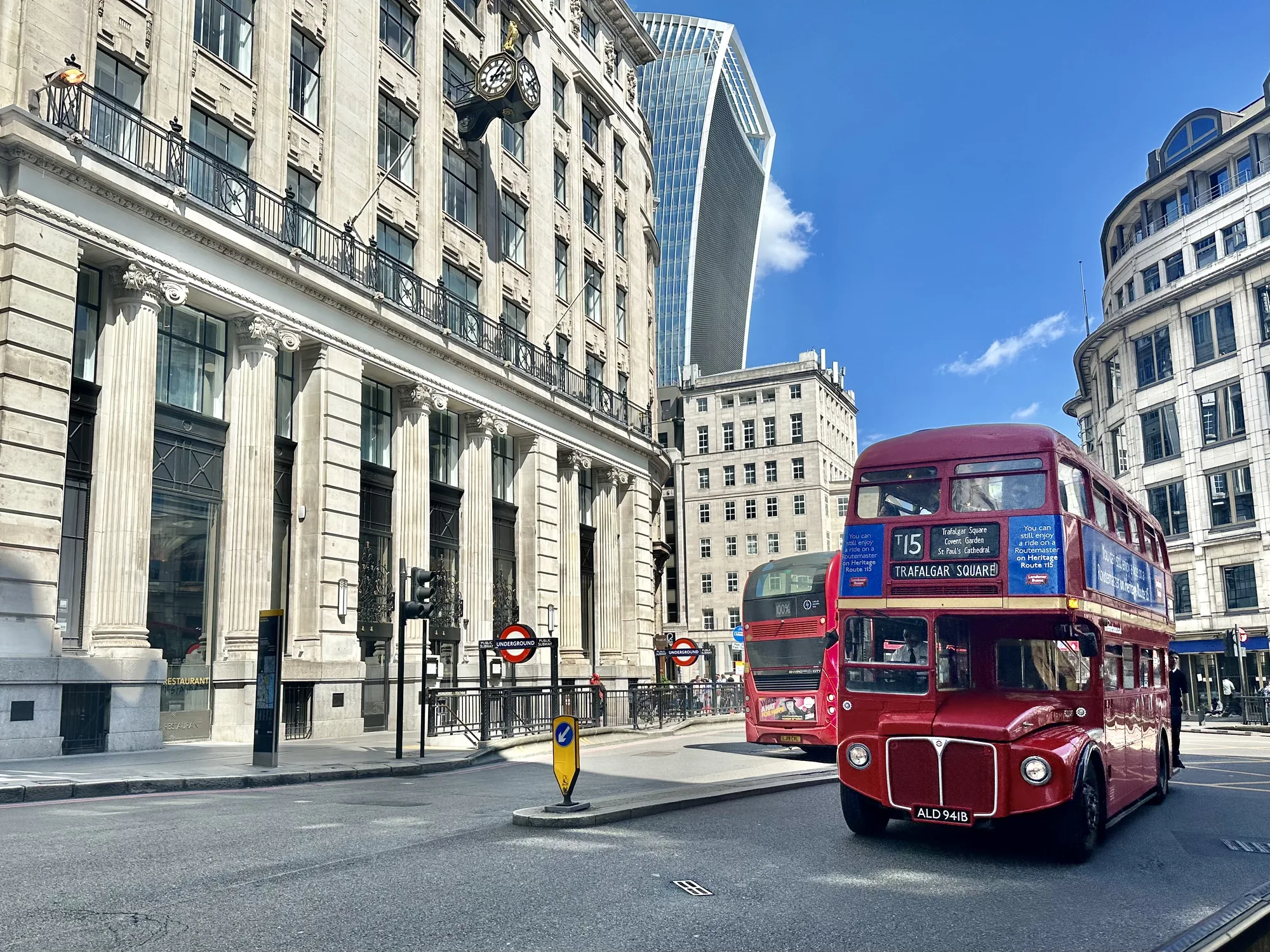 写真はイギリスのロンドンで撮影されたもので、晴れた日の街の様子が写っています。石造りの重厚な建物が並び、歴史を感じさせる景観です。左側の建物には大きな時計が設置されています。背景には、モダンな超高層ビル「ザ・ウォーキー・トーキー」が見えます。道路には、ロンドン名物の赤い二階建てバスが2台走っています。手前のバスには「TRAFALGAR SQUARE」と書かれており、トラファルガー広場方面に向かうルートであることがわかります。路面には地下鉄の入り口を示す標識があります。全体的に、ロンドンの新旧が融合した風景が印象的な写真です。