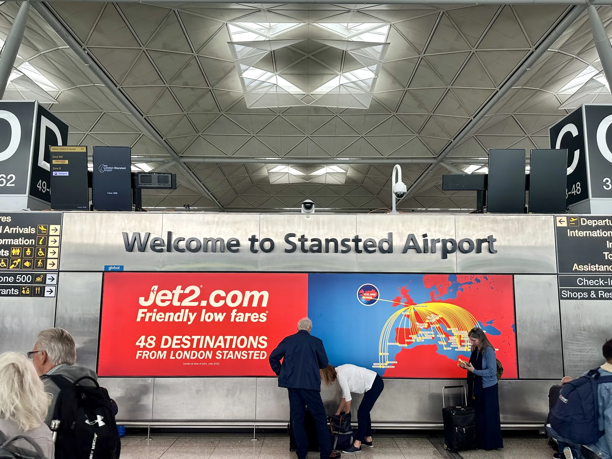 写真は、ロンドンのスタンステッド空港の内部を写しています。天井は特徴的な幾何学模様で構成されており、自然光を取り込む構造になっています。写真の中央には、空港名「Welcome to Stansted Airport」が掲示されています。その下には、航空会社Jet2.comの広告があり、「Friendly low fares（手頃な運賃）」というキャッチフレーズと、ロンドン・スタンステッド空港から48の目的地へのフライトがあることが示されています。広告の前には数人の旅行者がおり、スーツケースを整理したり、広告を見たりしています。右側には出発便の情報やショップ、レストランの案内を示す看板があります。写真全体は、空港の出発エリアの賑わいと旅行者の様子を捉えています。