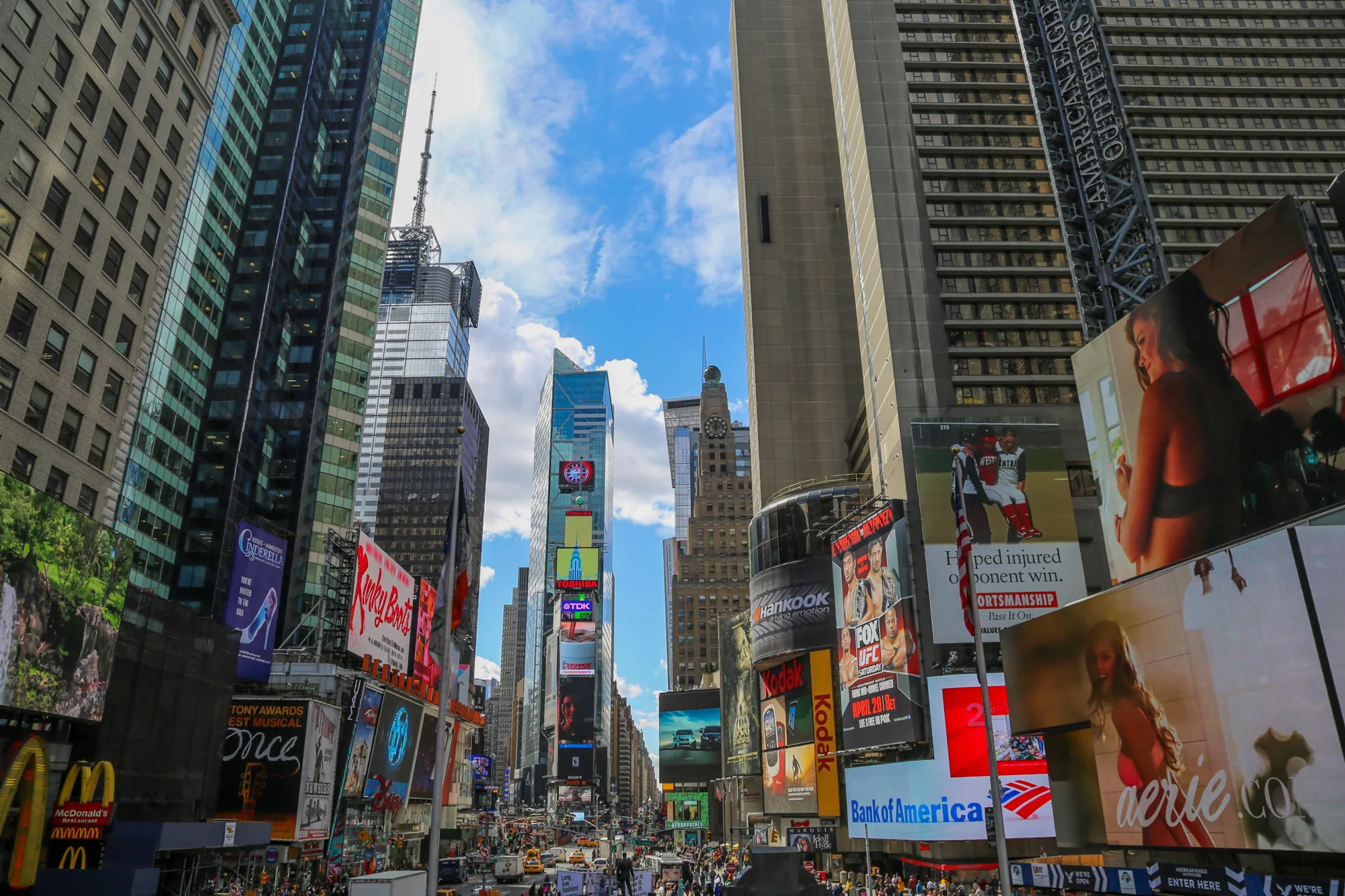 写真はアメリカ合衆国ニューヨークのタイムズスクエアを撮影したものです。高層ビルが立ち並び、その壁面には様々な広告看板が所狭しと掲げられています。青空が部分的に見え、ビル群の隙間から光が差し込んでいます。地上には多くの人々が行き交い、タクシーなどの車両も走っており、非常に賑やかな様子が伝わってきます。マクドナルドのロゴや、各種ブランドの広告、ミュージカルの広告など、多様な情報が視覚的に詰め込まれています。全体的に活気があり、エネルギッシュな都会の雰囲気が感じられます。