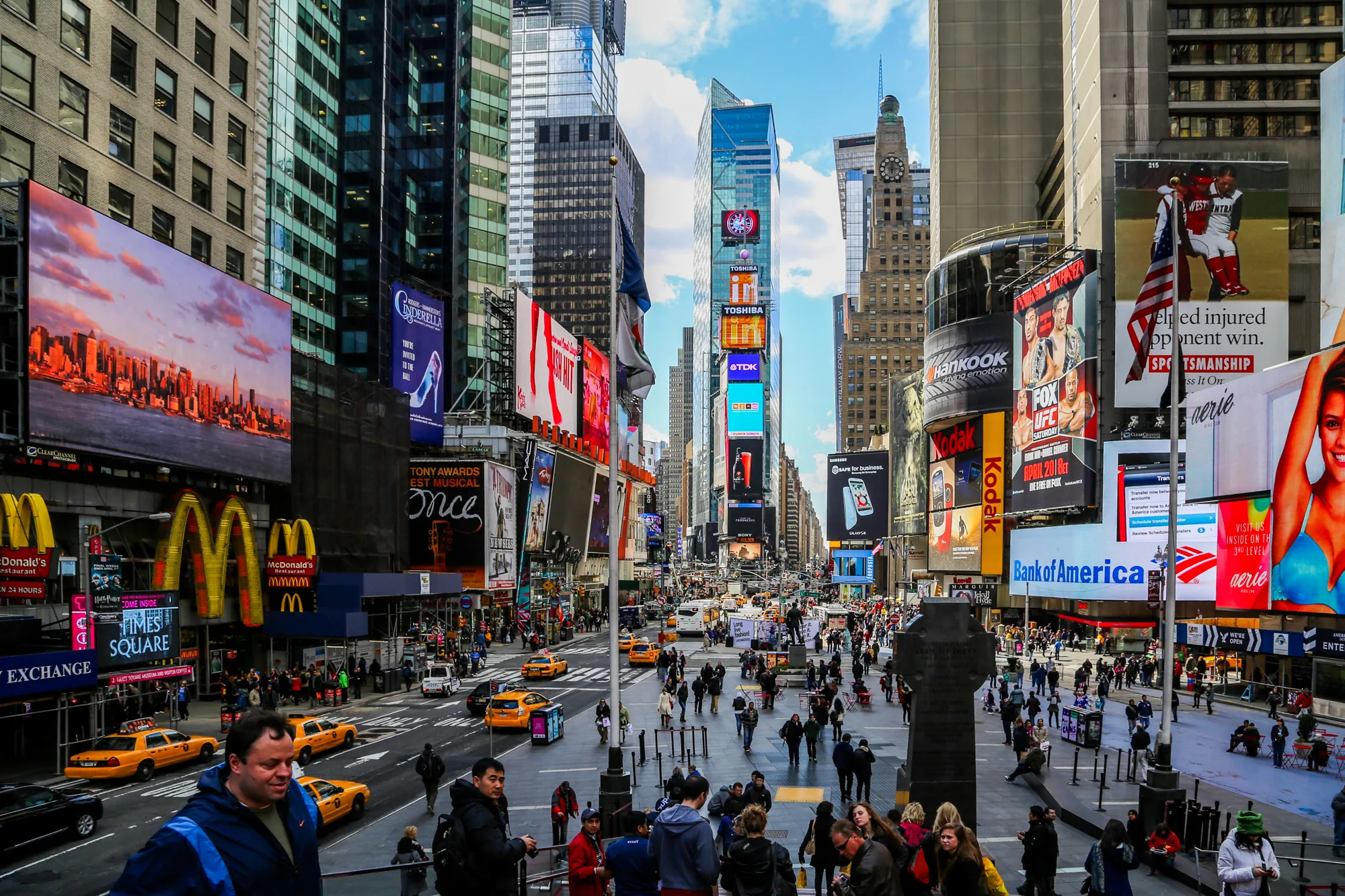 写真はニューヨークのタイムズスクエアを捉えたものです。高層ビルが立ち並び、それらを覆い隠すように巨大な広告看板が林立しています。看板には、マクドナルド、コダック、東芝、バンク・オブ・アメリカなど、様々な企業のロゴや広告が鮮やかに表示されています。道路には黄色いタクシーが走り、歩道や広場には大勢の人々が行き交い、賑わいを見せています。  写真全体は、エネルギーと活気に満ち溢れた雰囲気です。カラフルな広告、忙しく行き交う人々、高層ビルのコントラストが、ニューヨークの独特な都市景観を作り出しています。空は晴れており、わずかに雲が見えます。人々の服装は様々で、観光客や地元住民が混ざり合っているようです。写真全体から、タイムズスクエアの喧騒と刺激が伝わってきます。