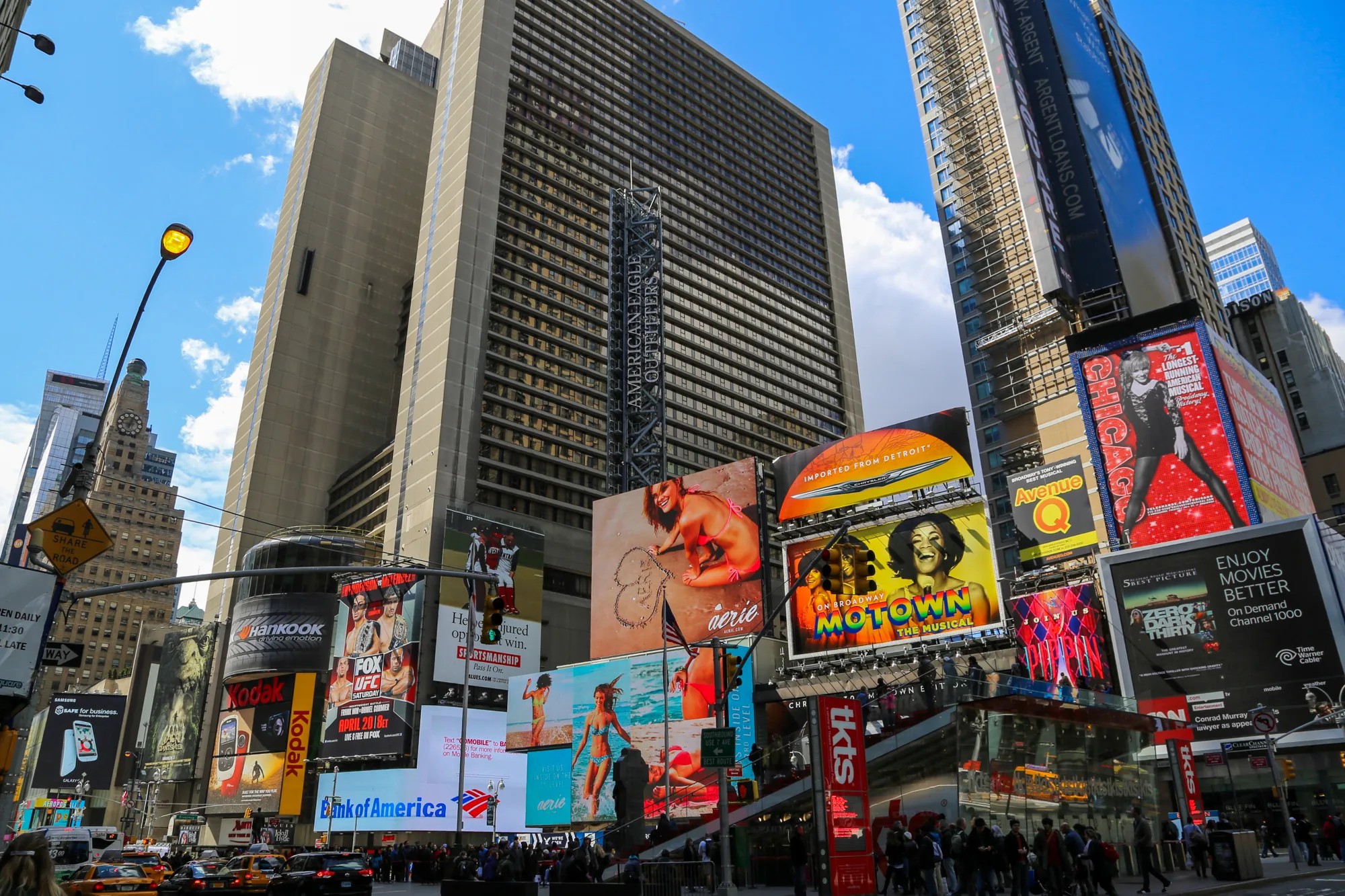 この写真はアメリカ、ニューヨークのタイムズスクエアを捉えたものです。画面全体に高層ビルがそびえ立ち、それらの壁面には様々な企業の巨大な広告看板が隙間なく設置されています。例えば、Hankook、Kodak、FOX UFC、Bank of America、American Eagle Outfitters、Aerie、Chrysler、Motown The Musical、Chicagoなどの広告が見られます。空は晴れており、一部に白い雲が浮かんでいます。  街路にはタクシーやバスが走り、多くの人々が行き交っており、非常に賑やかな様子が伝わってきます。歩行者用の信号機や標識も確認できます。写真全体から、タイムズスクエアの喧騒と活気が感じられます。