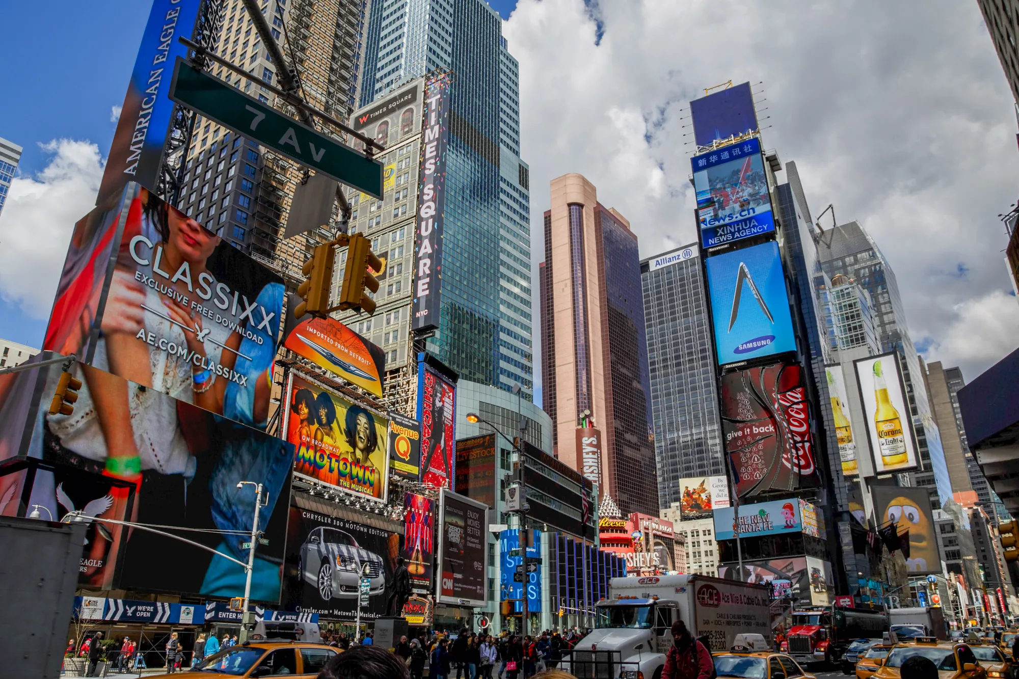 写真は、アメリカ合衆国ニューヨークのタイムズスクエアで撮影されたものです。高層ビルが立ち並び、巨大な広告看板が所狭しと並んでいる様子が写されています。看板には、ファッションブランド「AMERICAN EAGLE」、無料ダウンロードサイト「CLASSIXX」、ミュージカル「MOTOWN」、自動車「CHRYSLER 300」など様々な広告が表示されています。また、「TIMES SQUARE」と大きく書かれた看板や、中国の新華社通信の広告も見られます。道路には黄色いタクシーが走り、多くの人々が行き交っており、タイムズスクエアの活気ある雰囲気を伝えています。看板の光、車の光、自然光が混ざり合い、独特の色彩を作り出しています。全体的に、ニューヨークの象徴的な観光地であるタイムズスクエアの賑わいとエネルギーを感じさせる写真です。