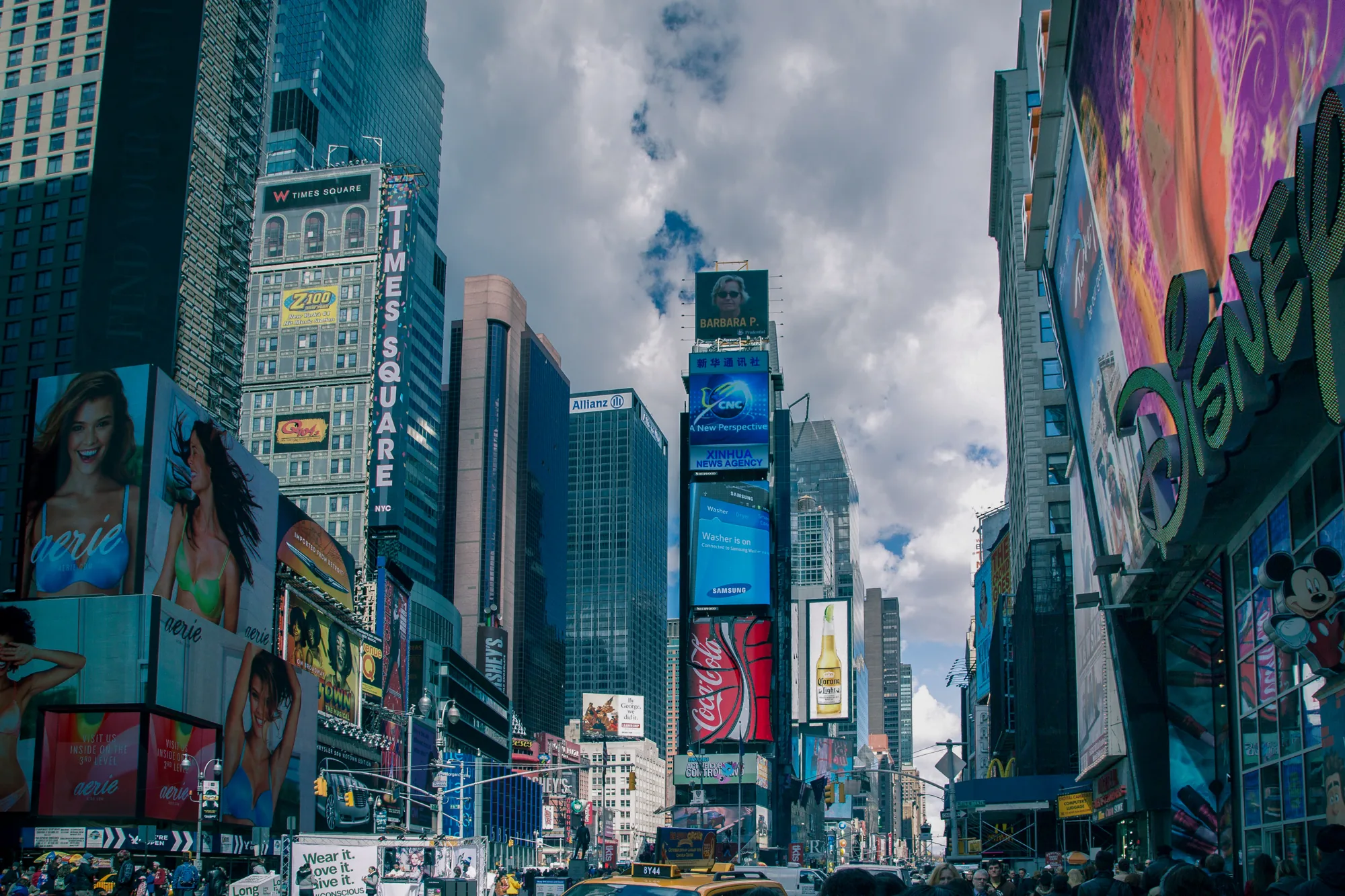 写真は、アメリカ合衆国ニューヨークのタイムズスクエアで撮影されたものです。画面全体に高層ビルが立ち並び、その壁面には巨大な広告が所狭しと並んでいます。広告は「aerie」「コカコーラ」「コロナ」などのブランドの他、ミュージカルの宣伝なども見られます。空は曇りがちですが、建物の隙間から少しだけ青空が見えています。画面下部には多くの人々が写っており、タイムズスクエアの賑わいを伝えています。黄色いタクシーが写りこんでおり、ニューヨークらしさを演出しています。ミッキーマウスの大きな看板も確認できます。全体的に鮮やかで活気のある雰囲気です。