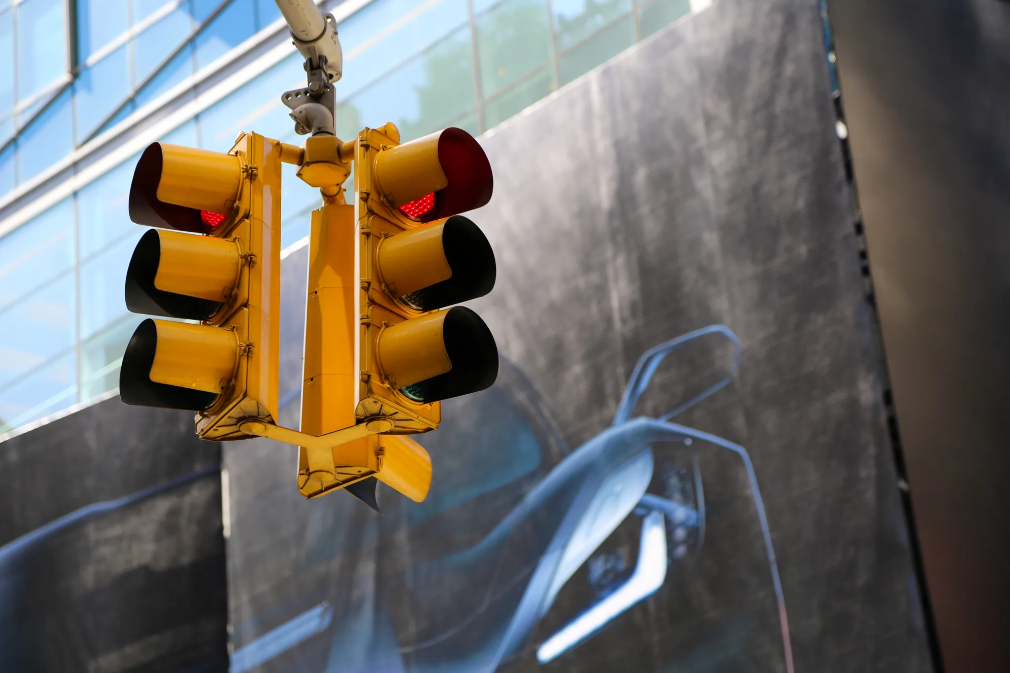 ニューヨークで撮影された、鮮やかな黄色の信号機を捉えた写真です。信号は全て赤色に点灯しており、一時停止を促しています。背景には高層ビルのガラス張りのファサードが一部見え、空の青色が映り込んでいます。また、巨大な広告看板のようなものがあり、抽象的な青色のデザインが施されています。信号機は金属製で、経年による汚れや傷みが見られますが、ニューヨークの都市景観の一部として存在感を示しています。全体的に、都会の喧騒と交通の制御を象徴するような、力強いイメージの写真です。