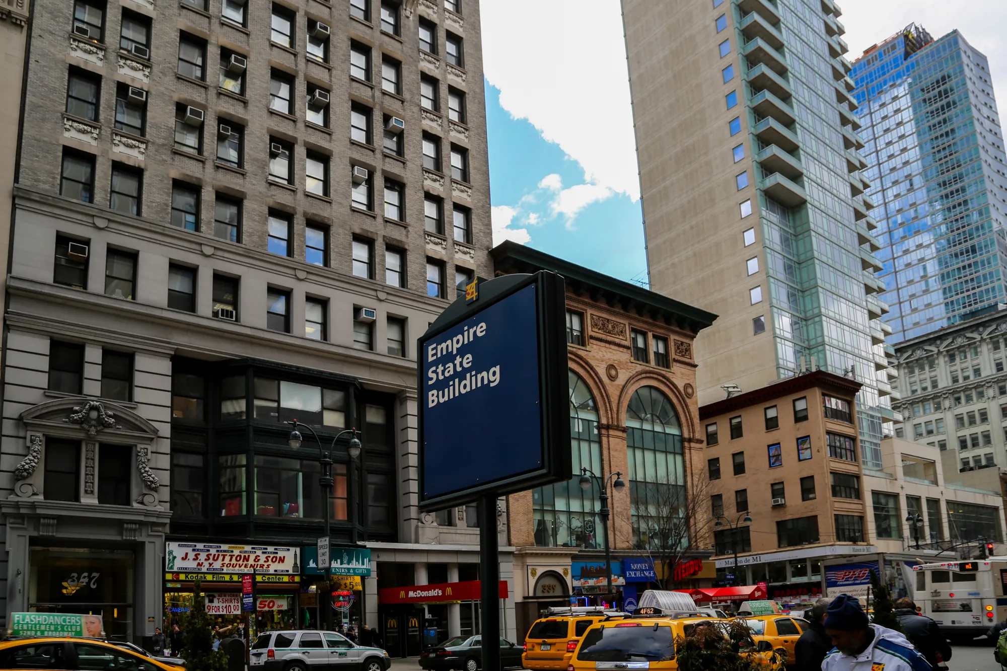 この写真はアメリカ合衆国、ニューヨーク市で撮影された都市景観です。画面の中心には「Empire State Building」と書かれた青い看板があり、エンパイアステートビルディングへの道のりを示唆しています。その奥には、様々な建築様式の建物が立ち並び、新旧の建物が混在している様子が伺えます。手前には黄色いタクシーが複数台停まっており、ニューヨークらしい賑やかな街の雰囲気を醸し出しています。また、写真にはマクドナルドの店舗や土産物店など、観光地らしい要素も確認できます。空には一部青空が見え、高層ビル群の合間から覗く空が印象的です。全体的にやや曇り空で、落ち着いた雰囲気の写真です。歩道には人が行き交い、都会の喧騒が感じられます。