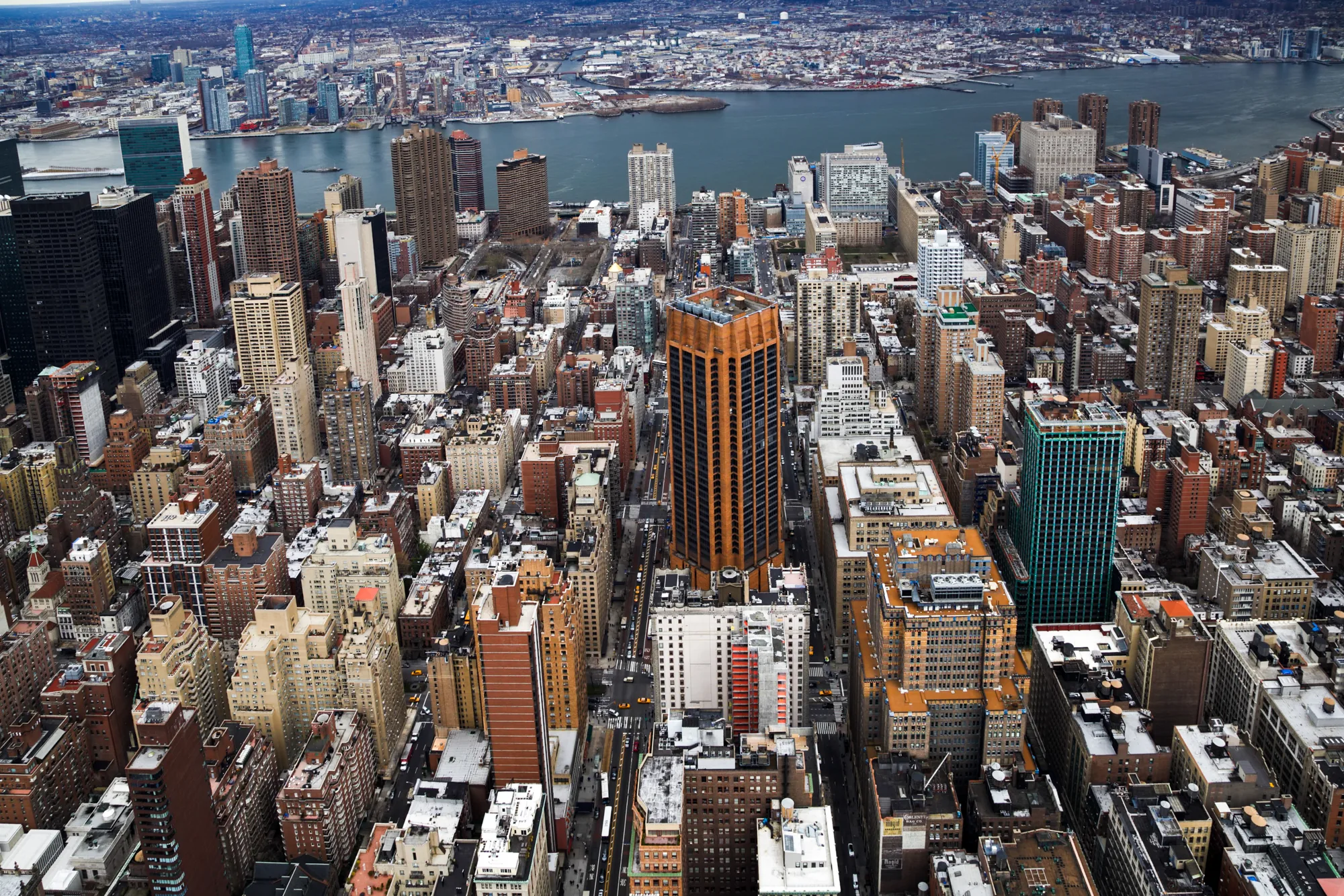 この写真は、アメリカ合衆国ニューヨーク市のマンハッタン地区を、高所から見下ろした航空写真です。画面全体に高層ビルが密集しており、様々な形や高さの建物がひしめき合っています。建物の色合いは、茶色、ベージュ、グレーなどが中心で、一部にオレンジ色や緑色の高層ビルも見られます。ビルの間には道路が走り、小さく車やタクシーが写っています。遠景には、イースト川やロングアイランドシティが見え、さらに遠くには霞んだ空が広がっています。全体的に、都会の賑わいと高層建築の圧倒的なスケール感が伝わってくる写真です。