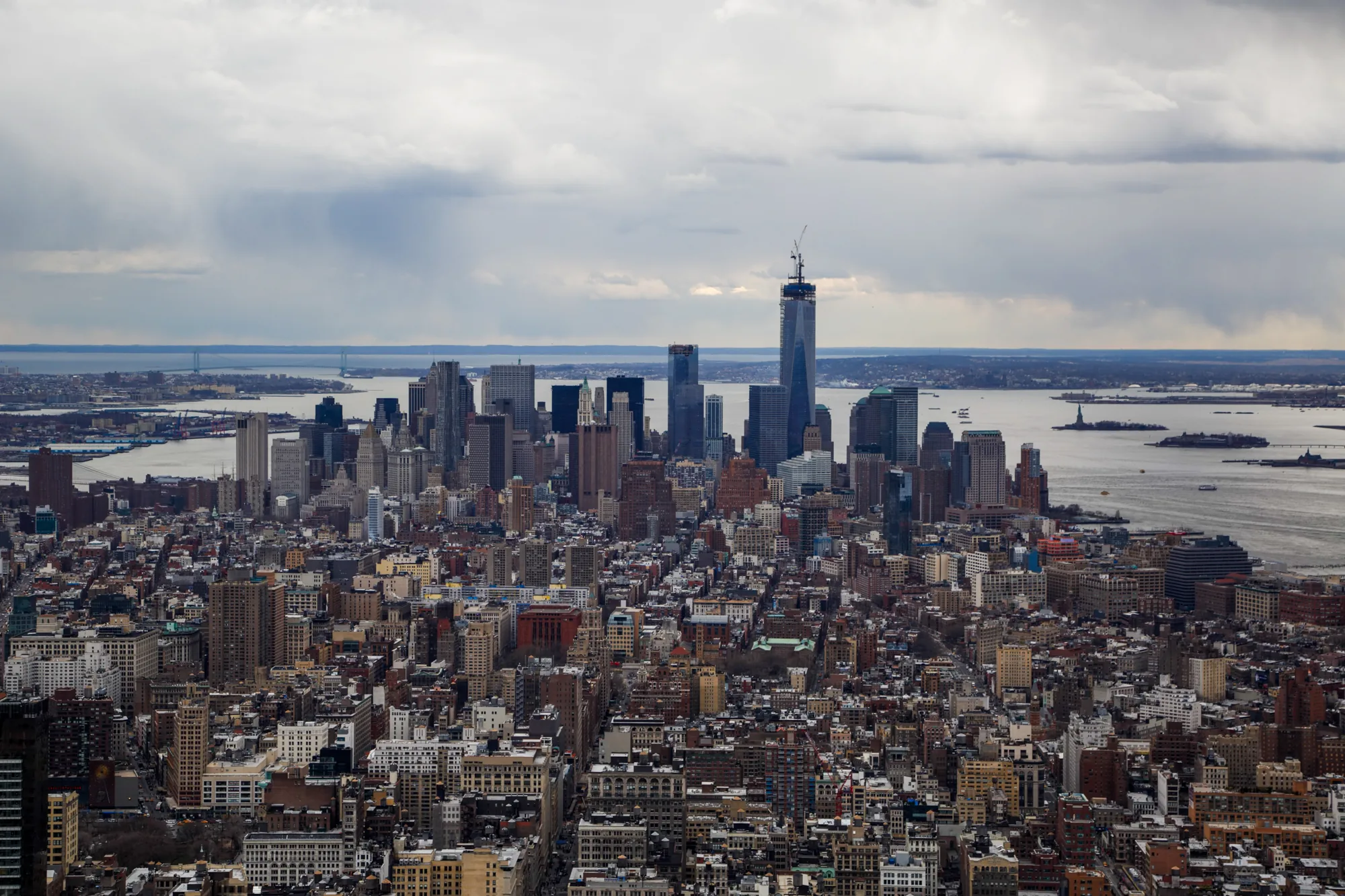 写真はアメリカ合衆国ニューヨーク市を上空から撮影したものです。手前には高層ビルが密集した市街地が広がり、遠景には高層ビル群のスカイライン、さらにその奥には海が見えます。画面中央には、特に高い高層ビルであるワンワールドトレードセンターがそびえ立っています。空は厚い雲に覆われており、全体的にどんよりとした天気であることがわかります。遠景の海には自由の女神像が小さく写っています。都市部は建物が密集しており、細い道路が網の目のように走っている様子がわかります。全体的にグレーがかった色調で、都会の雰囲気と少し物憂げな雰囲気が感じられます。