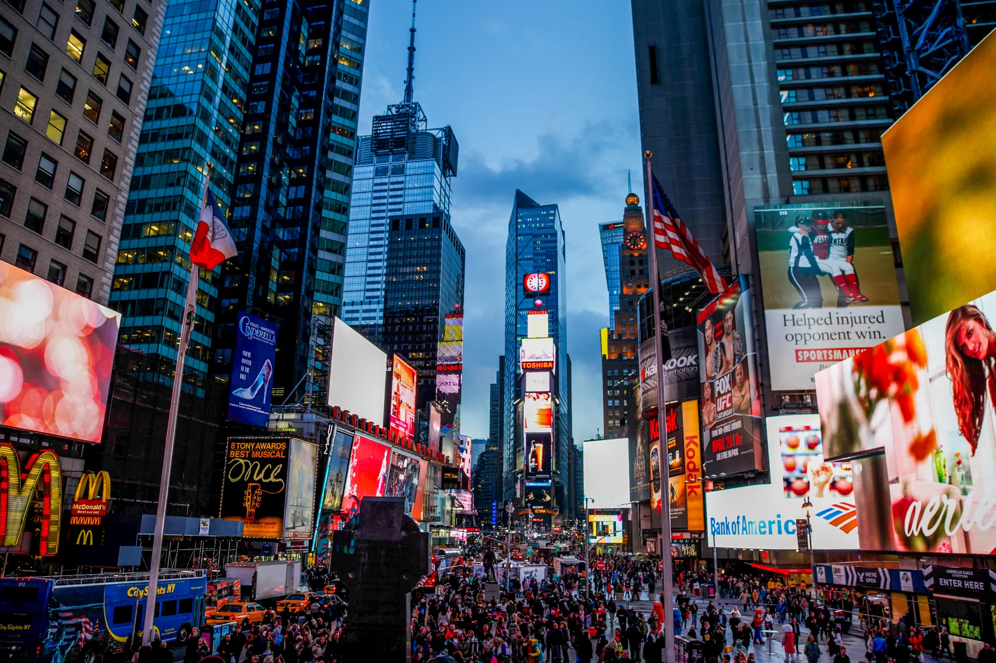 この写真はアメリカ合衆国ニューヨーク市のタイムズスクエアを捉えたものです。夕暮れ時の空の下、高層ビルが立ち並び、その壁面は巨大な広告看板で埋め尽くされています。マクドナルド、コダック、TOSHIBA、バンクオブアメリカなど、様々な企業のロゴや広告が鮮やかに輝いています。群衆が広場を埋め尽くし、活気に満ちた雰囲気が伝わってきます。アメリカ国旗やニューヨーク市の旗も掲げられています。道路にはタクシーや観光バスが走っており、街の喧騒を感じさせます。空はまだ明るく、ネオンサインの光と相まって幻想的な光景を作り出しています。
