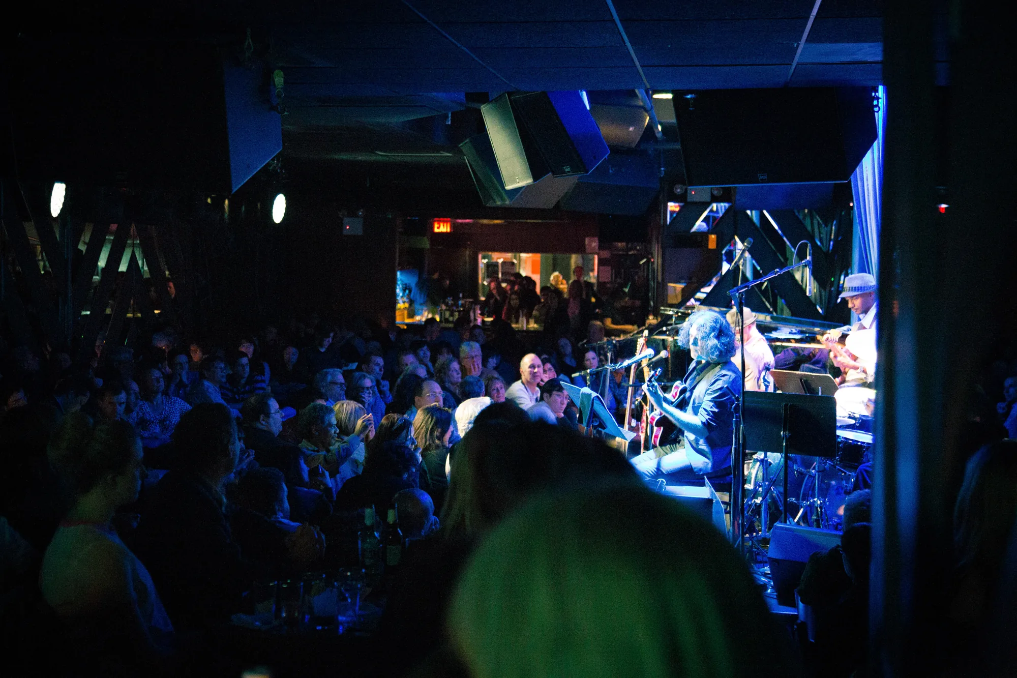 写真は、アメリカ・ニューヨークのライブハウスで撮影されたものです。会場内は薄暗く、青い照明が印象的です。ステージ上ではミュージシャンたちが演奏しており、ギターやキーボード、ドラムセットなどが見えます。観客席は人で埋め尽くされており、テーブルを囲んで音楽を楽しんでいる様子がうかがえます。天井にはスピーカーが設置されており、迫力のあるサウンドが期待できます。全体的に、親密で活気のあるライブイベントの雰囲気が伝わってきます。