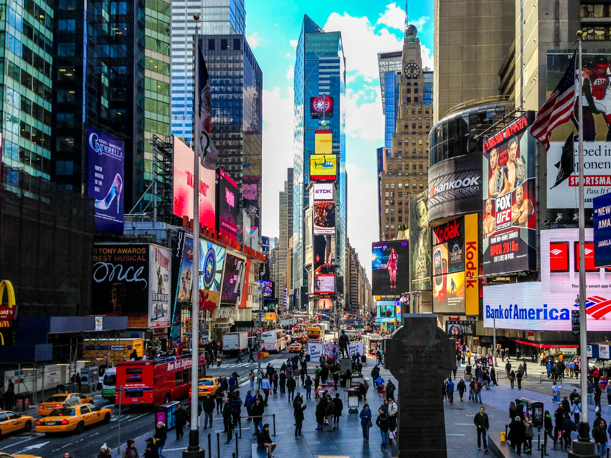写真はアメリカのニューヨーク、タイムズスクエアの様子を捉えています。高層ビル群が立ち並び、その壁面には巨大な広告が所狭しと掲示されています。ミュージカル「シンデレラ」や「ONCE」などの広告のほか、様々な企業広告が色鮮やかに輝いています。画面中央には多くの人々が行き交い、観光客や地元の人々で賑わっています。イエローキャブや観光バスが道路を走り、活気あふれる都会の風景を象徴しています。画面手前には像があり、多くの人がその周辺に集まっています。全体的に明るく鮮やかな色彩で、タイムズスクエアの華やかさを表現しています。