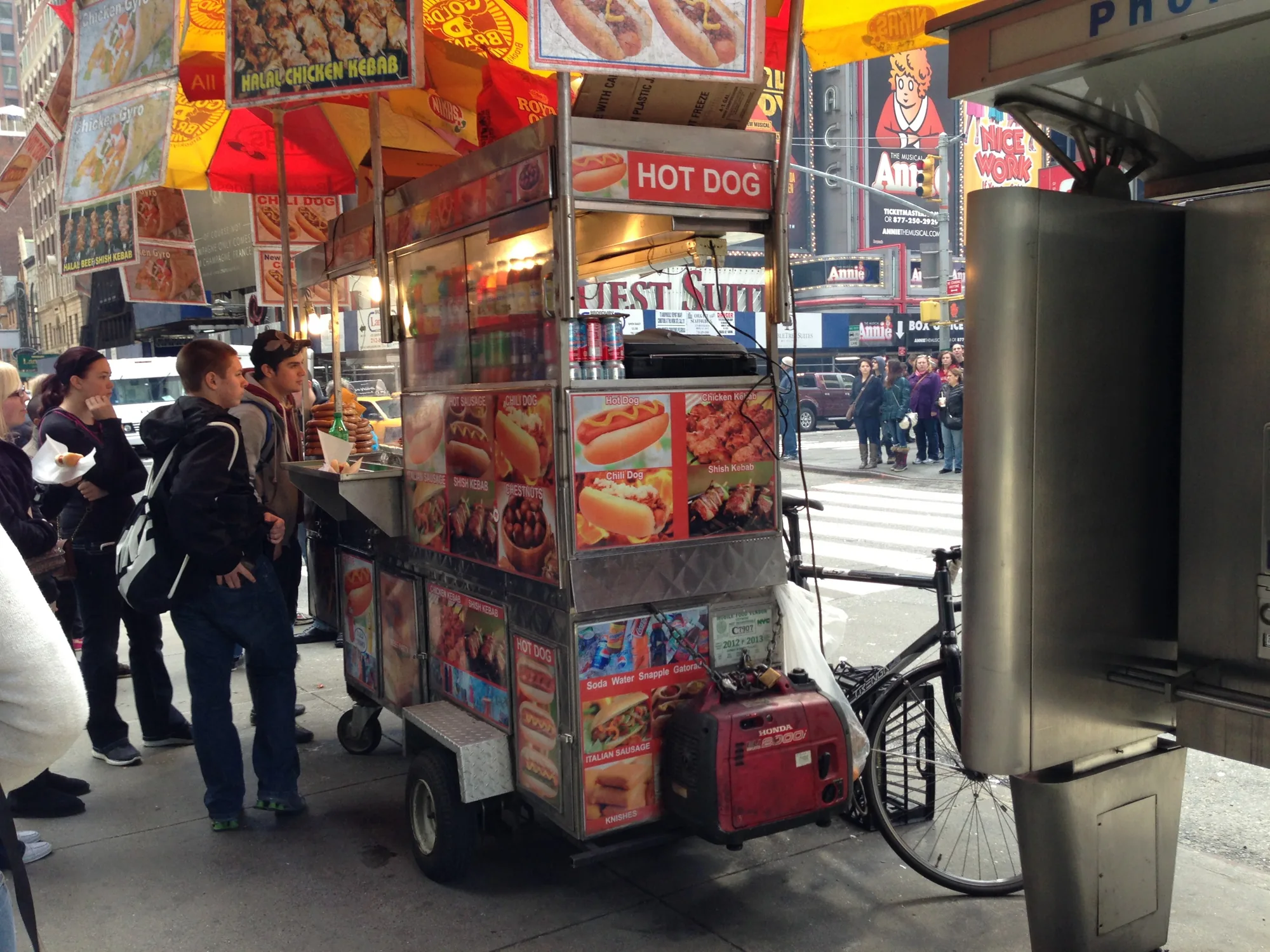 これはアメリカ、ニューヨークで撮影された写真です。賑やかな街並みの歩道に、ホットドッグやケバブを売る屋台が出ています。屋台のカラフルな看板には、ホットドッグやチキンケバブの写真が大きく載っており、食欲をそそります。数人の客が屋台に立ち寄り、食べ物を注文しているようです。背景にはタイムズスクエアのようなネオンサインが輝くビル群が見え、ニューヨークの活気ある雰囲気が伝わってきます。屋台の脇には自転車が停められており、街の風景の一部となっています。写真全体を通して、ニューヨークの日常的な風景がリアルに切り取られています。