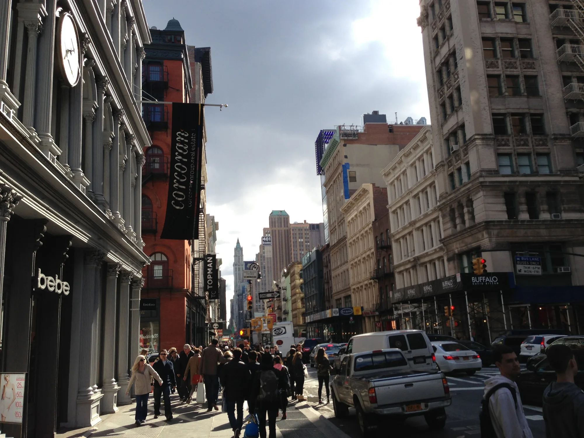 この写真はアメリカ、ニューヨークの街並みを捉えたものです。通りの両側には高層ビルが立ち並び、歩道には多くの人が行き交っています。左側の建物には「bebe」というショップがあり、その隣には「Corcoran」という不動産会社の旗が掲げられています。遠くには尖塔のある建物が見え、ニューヨークらしい景観を形成しています。空は少し曇っており、通りの奥は明るく光が差し込んでいます。車やトラックが道路を走り、交通量も多いことがわかります。全体的に活気があり、都会的な雰囲気の写真です。
