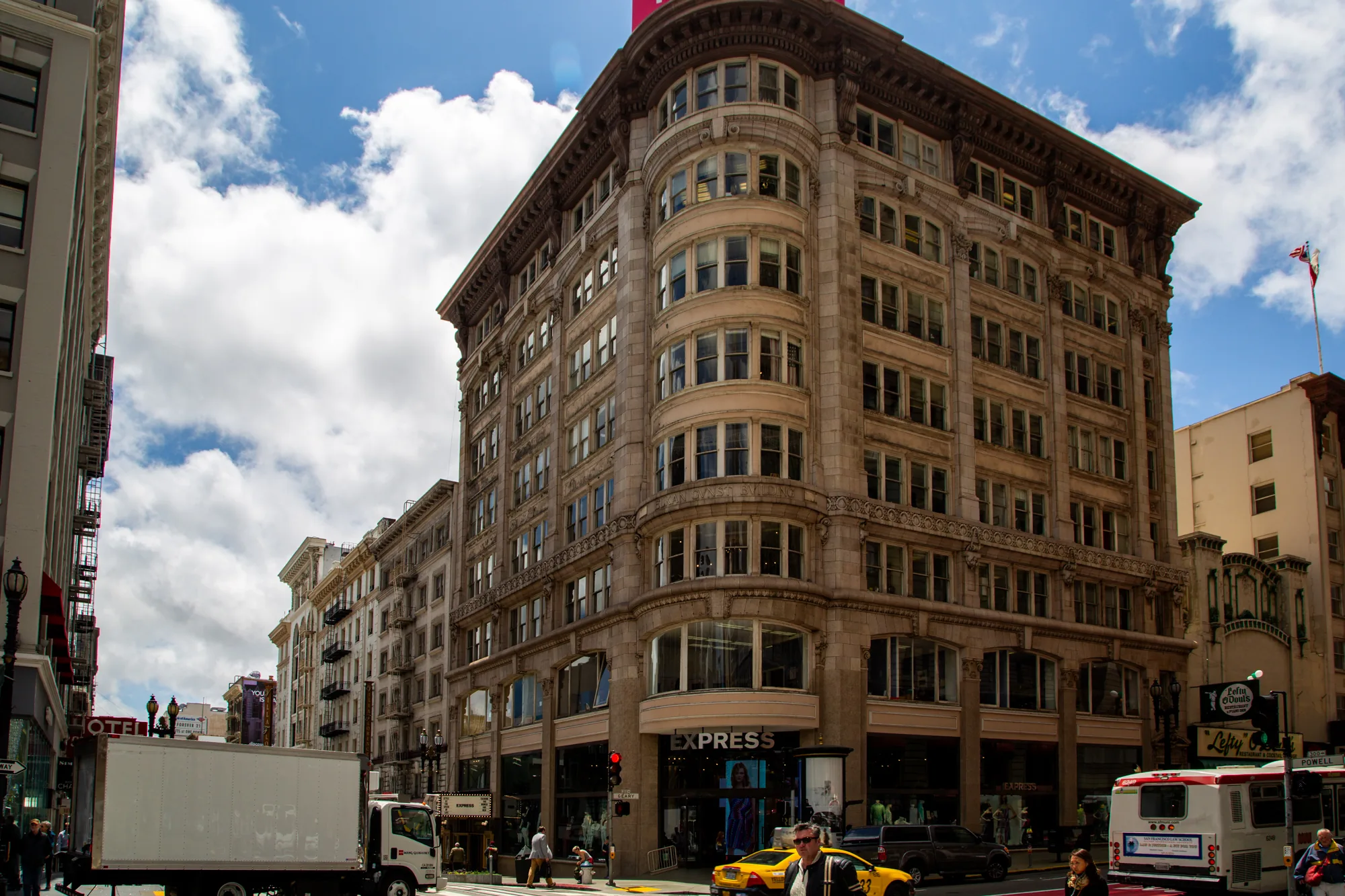 写真は、アメリカのサンフランシスコで撮影された街並みの様子を捉えています。空は青く、白い雲が浮かんでおり、日差しが差し込んでいます。画面中央には、石造りの重厚な建物がそびえ立っており、クラシックな建築様式が特徴的です。建物の1階には「EXPRESS」というアパレルショップがあり、多くの人が行き来しています。  道路には、白いトラックや黄色いタクシー、路線バスが走っており、活気のある都市の風景が表現されています。歩道には人々が歩いており、様々な人種や服装の人が見られます。画面右側には「Lefty O'Douls」というレストランの看板が見え、サンフランシスコらしい雰囲気を醸し出しています。写真全体から、サンフランシスコの賑やかさと歴史的な魅力が伝わってきます。