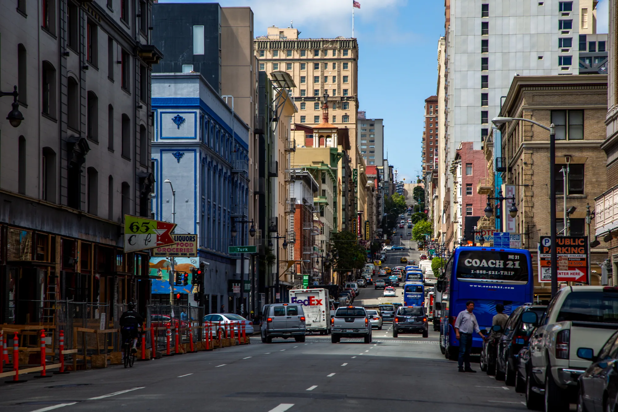 写真はアメリカのサンフランシスコで撮影されたものです。街の中心部の通りを捉えたもので、奥に向かって緩やかな坂道になっています。道路は交通量が多く、複数の車両が走行しています。手前には紺色の観光バスが停車しており、運転手らしき人物が立っています。道路の左右には建物が立ち並び、古いレンガ造りの建物や、近代的な高層ビルなどが混在しています。青空が広がり、日差しが差し込んでいますが、建物によって影ができ、明暗のコントラストが強調されています。通りの名前を示す「Turk」という標識が見えます。左手には工事現場があり、オレンジ色のフェンスが設置されています。全体的に活気があり、都会的な雰囲気の写真です。