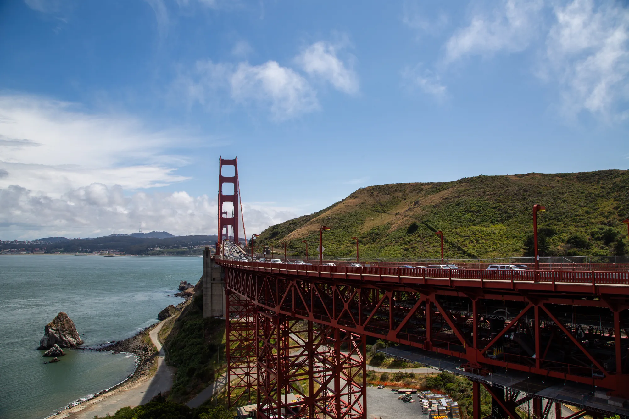 この写真は、アメリカ合衆国サンフランシスコの象徴である金門橋（ゴールデンゲートブリッジ）を捉えたものです。明るい青空と白い雲が広がる背景に、鮮やかな赤色の橋が堂々とそびえ立っています。橋の上を走る車の列から、橋が現在も活発に使われていることがわかります。橋の右側には緑豊かな丘が広がり、自然との調和が感じられます。橋の左側には青い海が広がり、岩礁が点在しています。写真全体から、サンフランシスコの美しい景観と、金門橋の存在感が伝わってきます。