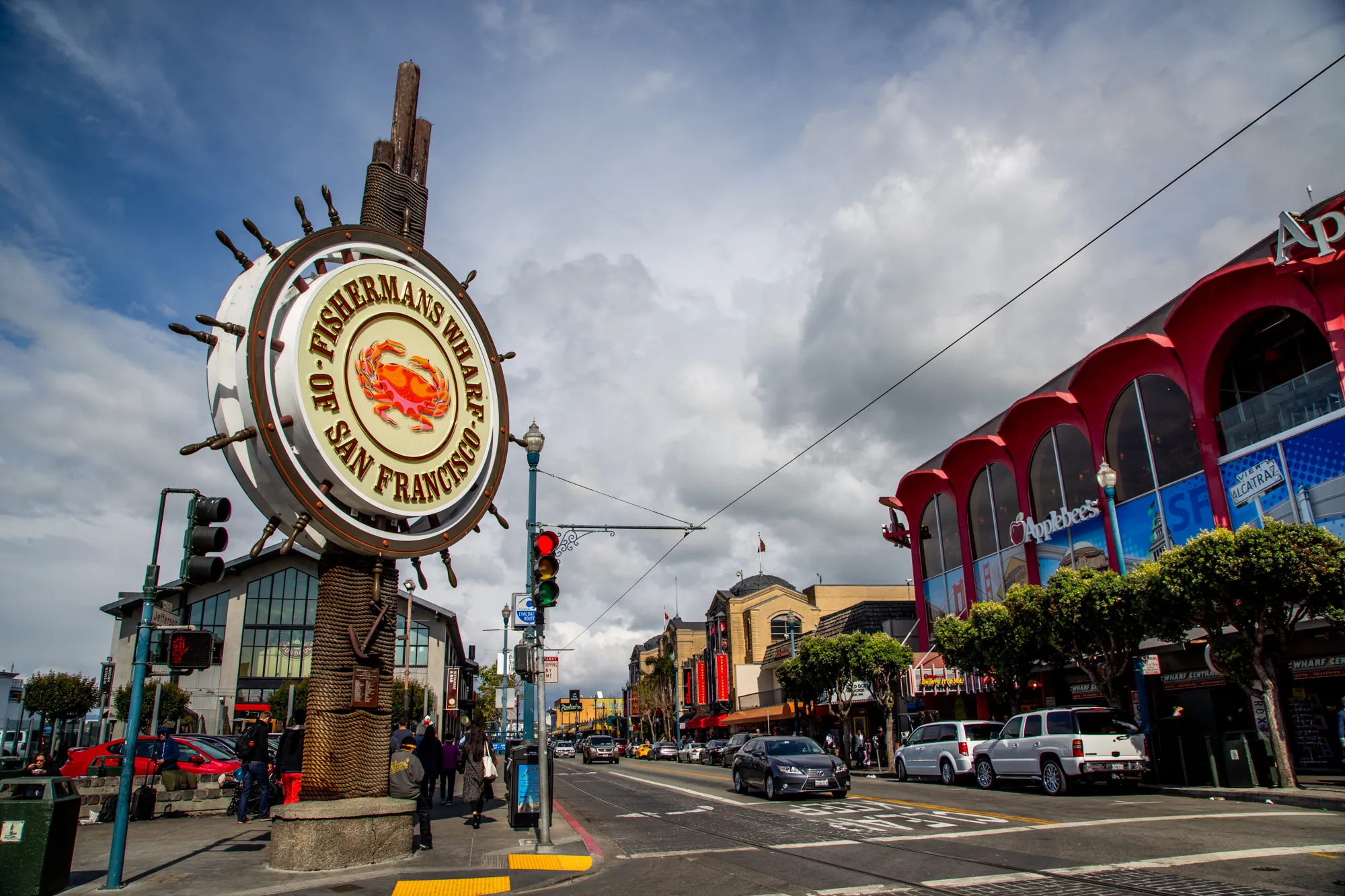 この写真はアメリカ、サンフランシスコの街並みを捉えたものです。手前には「FISHERMAN'S WHARF OF SAN FRANCISCO」と書かれた、カニのイラストが描かれた大きな看板があります。この看板は舵輪のような形状で、ロープで巻かれた柱の上に設置されています。看板の左には信号機と、歩行者が数人写っています。  道の向かい側には様々な建物が並んでいます。右側には赤いアーチ型の窓が特徴的な建物が見え、Applebee'sの看板が出ています。この建物の窓にはアルカトラズ島の広告が見えます。道路には車が数台走行しており、駐車車両も見られます。空は雲で覆われていますが、所々に青空も見えています。全体的に活気のある雰囲気の写真です。