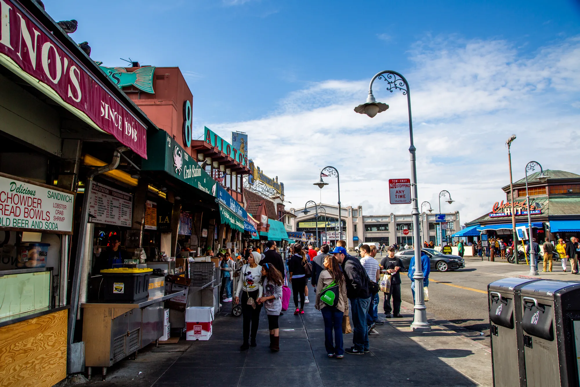 写真はアメリカのサンフランシスコ、おそらくフィッシャーマンズワーフ周辺の賑やかな通りを捉えています。青い空に白い雲が浮かび、明るい日差しが降り注いでいます。通り沿いには、シーフードレストランや土産物店などが軒を連ね、カラフルな看板や装飾が目を引きます。「NINO'S SINCE 1918」と書かれた看板や、「Delicious CHOWDER BREAD BOWLS」といったメニューが掲げられた店が見られます。  人々は行き交い、観光客が多い様子が伺えます。グループで歩いたり、店を覗いたり、会話を楽しんだりする姿が自然な雰囲気で写っています。通りの先には建物が見え、遠くまで視界が開けています。街灯や標識も設置されており、都会的な風景です。全体的に活気があり、観光地の賑わいを伝える写真です。