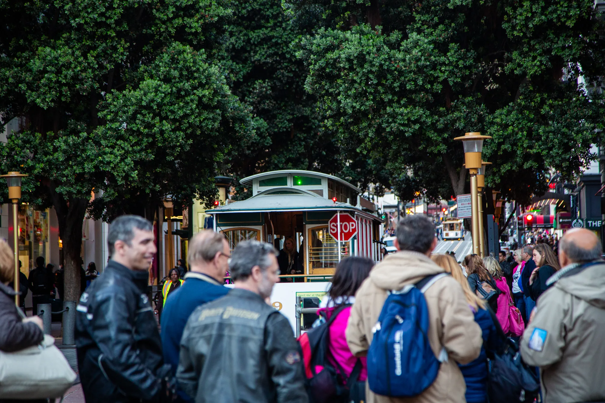 写真はサンフランシスコで撮影されたと思われる、街の風景を捉えたものです。焦点は、緑色の屋根を持つクラシックなケーブルカーに当てられています。ケーブルカーの前面には赤い「STOP」の標識が見え、その内部には乗客らしき人物がいます。  ケーブルカーの周囲には多くの人々がいて、列を作って待っているように見えます。人々は様々な服装をしており、バックパックを背負っている人もいます。背景には、街灯、道路標識、そしていくつかの建物が確認できます。木々が画面の上部を覆い、全体的に賑やかな街の雰囲気を醸し出しています。