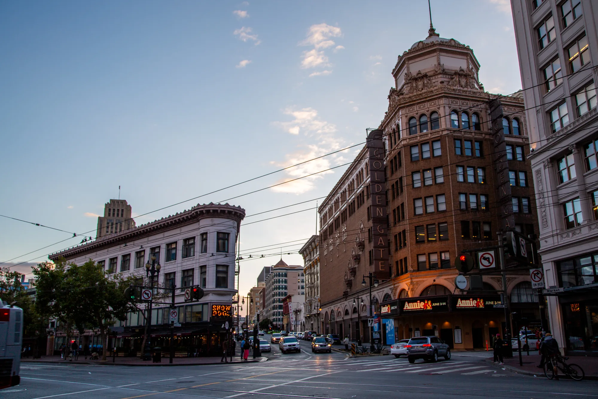 写真はアメリカのサンフランシスコで撮影された街の風景です。夕暮れ時の空が背景に広がり、建物群がシルエットのように浮かび上がっています。道路には車が複数走っており、交差点には歩行者や自転車に乗った人が見えます。左側の建物には「SHOW DOGS」と書かれたネオンサインが輝き、右側の建物には「GOLDEN GATE」と垂直に書かれた文字が見えます。また、「Annie」と書かれた劇場のような場所も写っています。電線が空を横切り、都市の景観を形作っています。全体的に、活気のある都市の夕暮れ時の様子が表現されています。