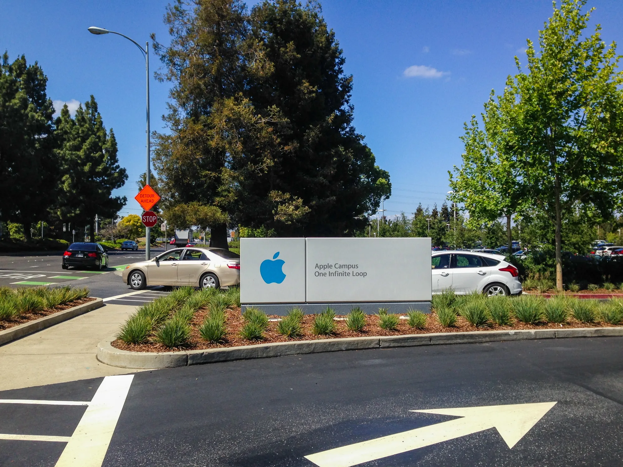 写真は、アメリカのサンフランシスコ近郊にあるApple Campusの入り口付近を撮影したものです。正面には、白い大きな看板があり、青色のAppleのロゴと「Apple Campus One Infinite Loop」という文字が書かれています。看板の周りには、緑の草が植えられ、整備された印象を受けます。  看板の背景には、緑豊かな木々が茂っており、青空が広がっています。道路には矢印のマークが描かれており、車の進行方向を示しています。交差点には、一時停止の標識や工事中の迂回路を示す標識も見られます。車も複数台写っており、Apple Campus周辺の日常の風景を捉えた写真です。全体的に明るい陽射しが降り注ぎ、穏やかな雰囲気です。