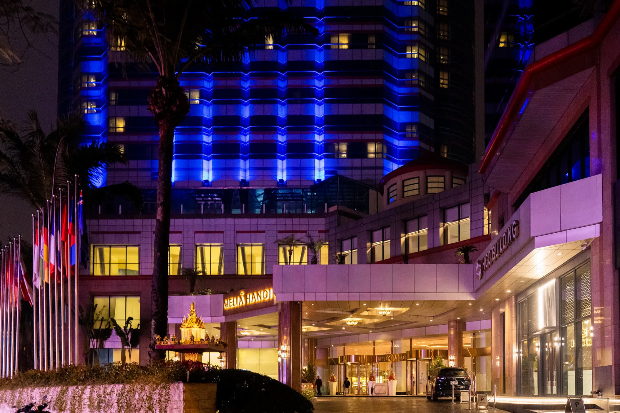 ベトナムのハノイで撮影された夜景写真です。メインは、ホテル「MELIA HANOI」の建物正面入口で、夜のライトアップが印象的です。高層部分は青色のライトで照らされており、モダンな雰囲気を醸し出しています。低層部分は暖色の照明で照らされ、落ち着いた雰囲気を演出しています。  ホテルの入口には、いくつかの旗が並んでおり、国際的な雰囲気を醸し出しています。入口の前には植栽があり、その中には東南アジア風の小さな祠のようなものが見えます。入口のロータリーには車が停まっており、ホテルを利用する人々の姿も見られます。写真全体から、ハノイの高級ホテルの夜の賑わいを感じ取ることができます。