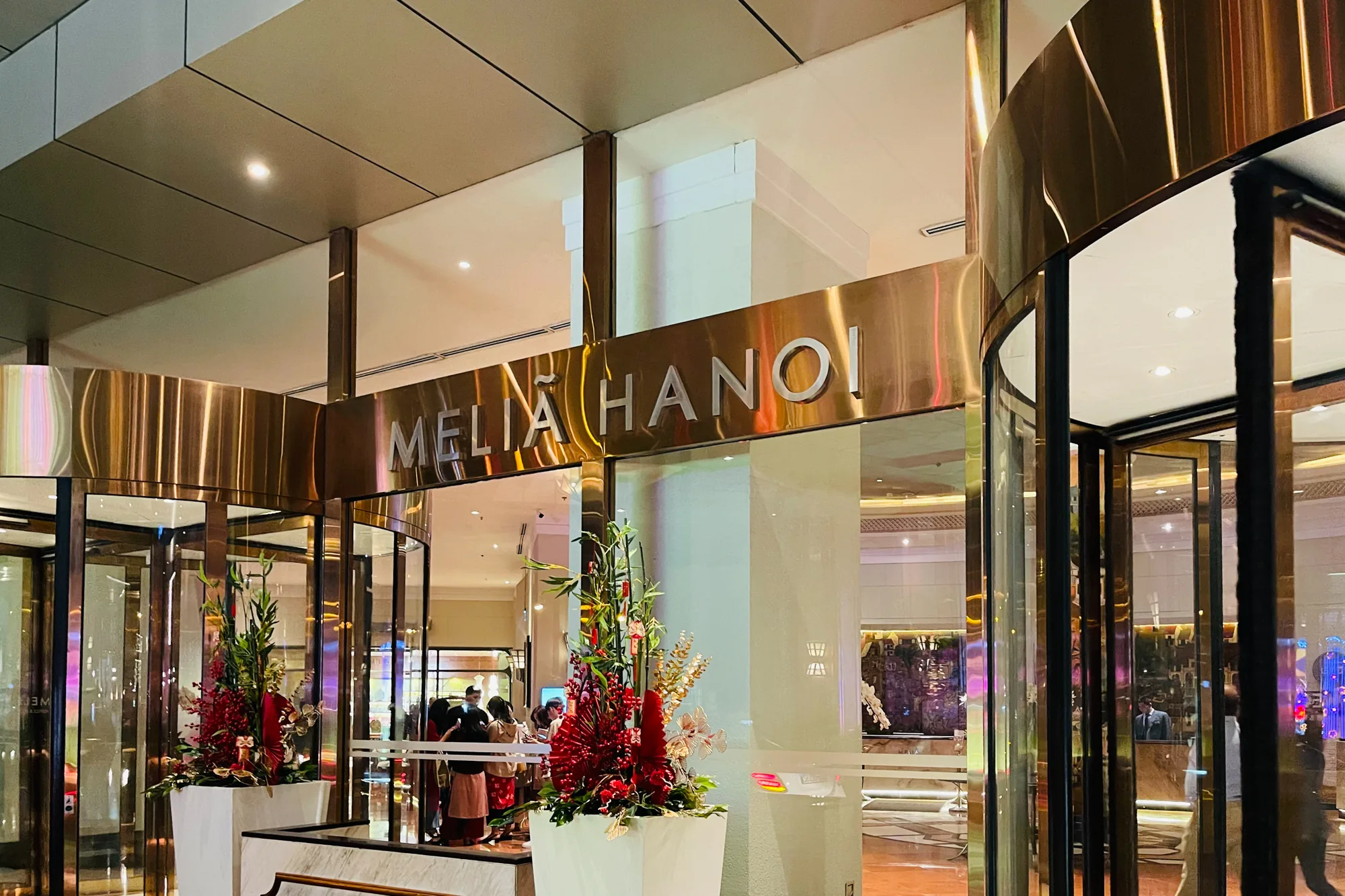 写真はベトナムのハノイにある「Melia Hanoi」ホテルの入口を写したものです。回転ドアの上には光沢のある金色の看板があり、白抜きの文字で「MELIA HANOI」とホテル名が示されています。入口の両脇には白いプランターに活けられた、赤を基調とした豪華なアレンジメントフラワーが飾られています。回転ドアの向こうには、ホテルのロビーが見え、人々が談笑している様子が伺えます。ロビーの奥には、カジノのような電飾が確認できます。全体的に上品で落ち着いた雰囲気であり、高級感を醸し出しています。