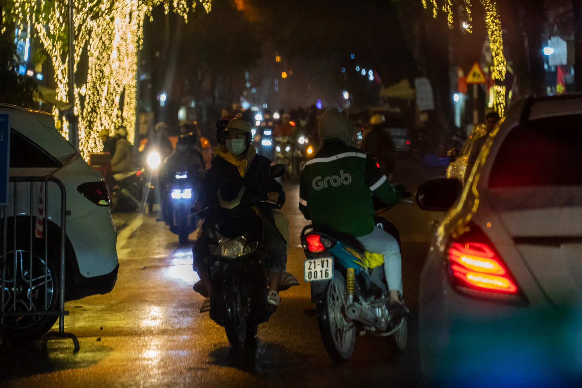 この写真はベトナムのハノイで撮影された夜の街の様子を捉えています。画面中央には、バイクに乗った人々が複数写っており、そのうちの一人は緑色のGrabのロゴが入ったジャケットを着ています。Grabは東南アジアで展開する配車サービスです。バイクのナンバープレートには「21-V1 0016」と記されています。  路面は濡れており、ライトが反射して光沢を帯びています。街路樹はイルミネーションで飾られており、背景の光とともに幻想的な雰囲気を醸し出しています。遠くには多くのバイクのヘッドライトが見え、交通量の多さを感じさせます。  画面の手前には、車のテールランプが赤く光っており、路上駐車している様子がうかがえます。全体的に薄暗い写真ですが、ライトやイルミネーションの光が人々の活動を照らし出しており、ハノイの夜の賑わいを表現しています。