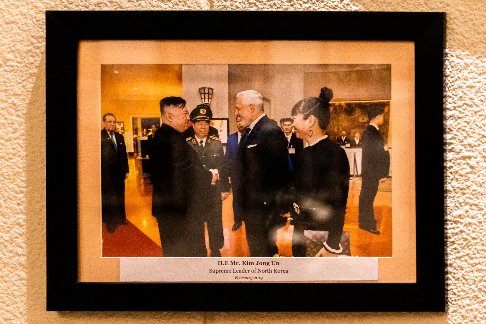 ベトナムのハノイで撮影された写真で、壁に飾られた額縁に入っています。額縁は黒色で、写真の周りにベージュ色のマットがあります。写真には、北朝鮮の最高指導者、金正恩氏が写っています。金正恩氏は、他の男性と握手を交わしています。その横には軍服を着た男性も写っています。その横には女性がおり、笑顔で会話をしているようです。写真の下には、「H.E Mr. Kim Jong Un, Supreme Leader of North Korea, February 2019」と記載された白いラベルが貼られています。写真全体は、暖色系の照明で照らされており、歴史的な瞬間を捉えた記録写真のような印象を与えます。