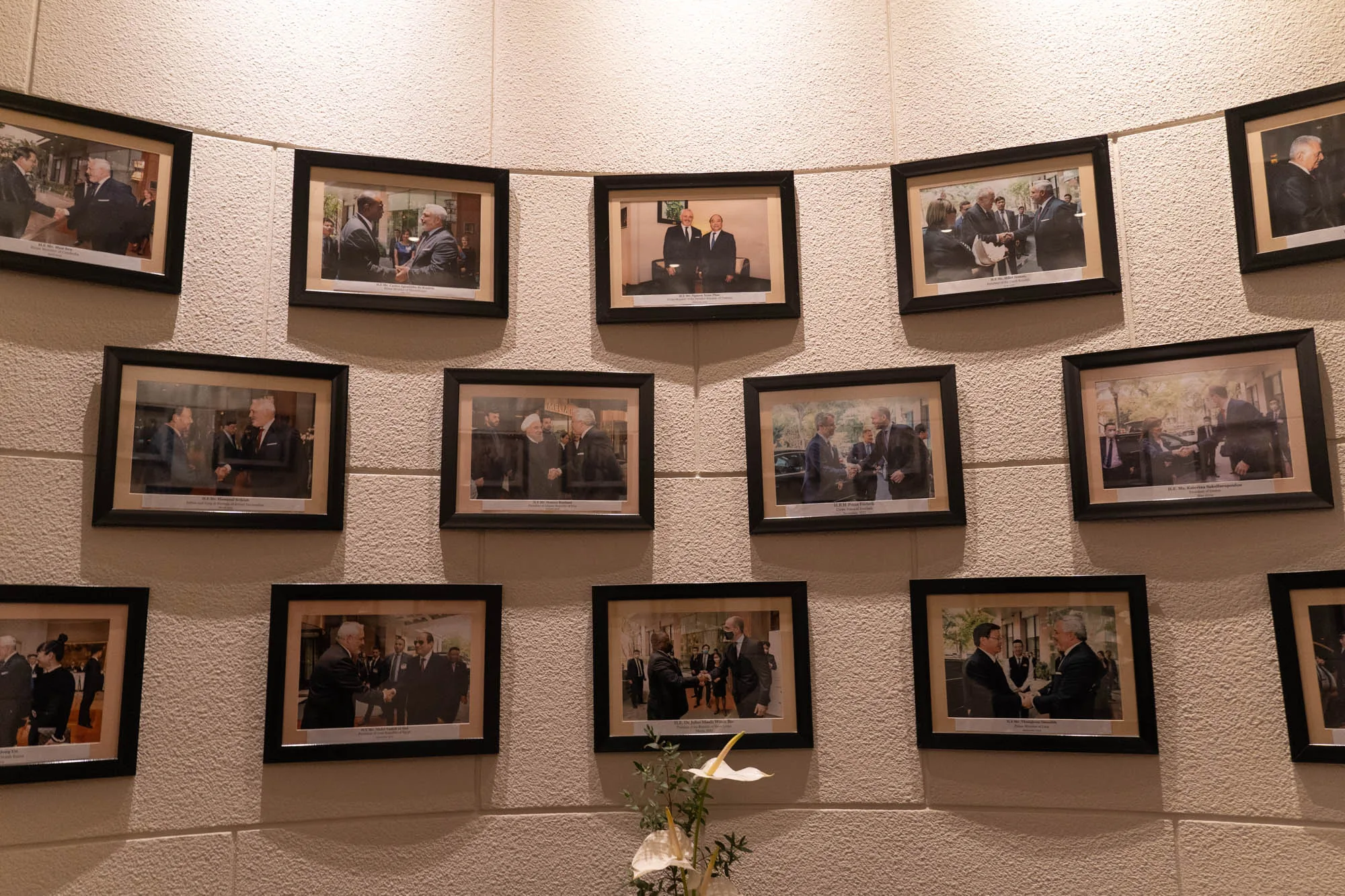 写真には、ハノイの壁に飾られた複数の写真が写っています。写真は黒いフレームに入れられており、壁は白い石膏のようなテクスチャを持っています。写真の内容は様々で、主にスーツを着た複数の人物が握手をしている場面が写っています。各写真の下には短い説明書きが見られます。壁の下部には、白いカラーと緑の葉を持つ花瓶が置かれています。壁の上部からは照明が当てられており、全体的に落ち着いた雰囲気の写真です。