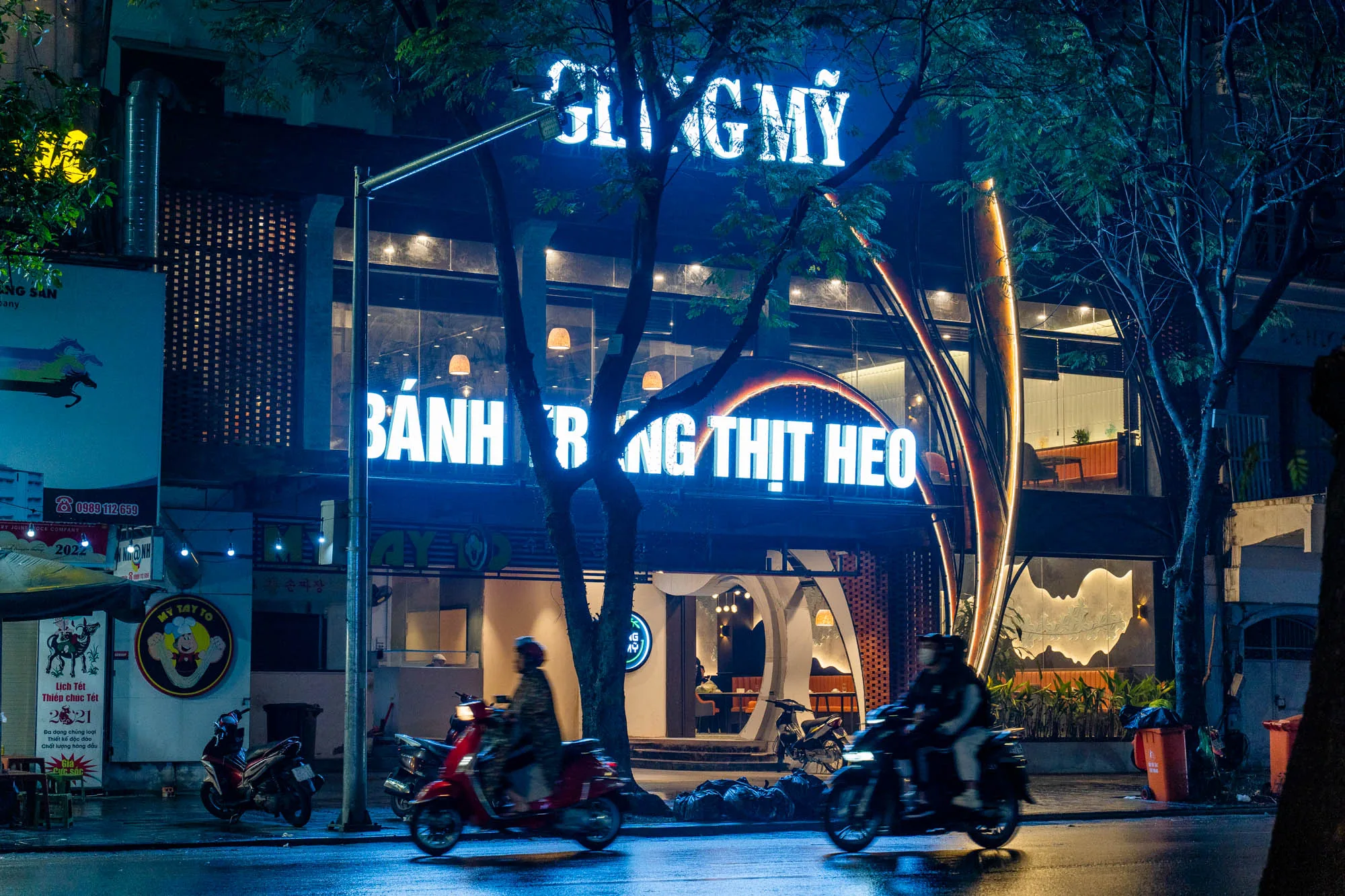 この写真はベトナムのハノイで撮影された夜の街並みを捉えています。前景には濡れた路面が広がり、二台のバイクが走行している様子がブレて写っています。画面中央には、大きな青色のネオンサインで店名と思われる文字「GLINGMY」と「BÁNH BÓNG THỊT HEO」が輝いています。このネオンサインは、おそらく店のもので、現代的なデザインのレストランかカフェのようです。店内には照明が灯り、テーブルや椅子が見えます。  左側には、看板や広告がいくつか掲示されており、ベトナム語の文字やイラストが描かれています。また、街路樹がフレームの両側に配置され、夜の街に独特の雰囲気を加えています。全体的に、湿った路面とネオンの光が、ハノイの活気ある夜の雰囲気を表現している写真です。