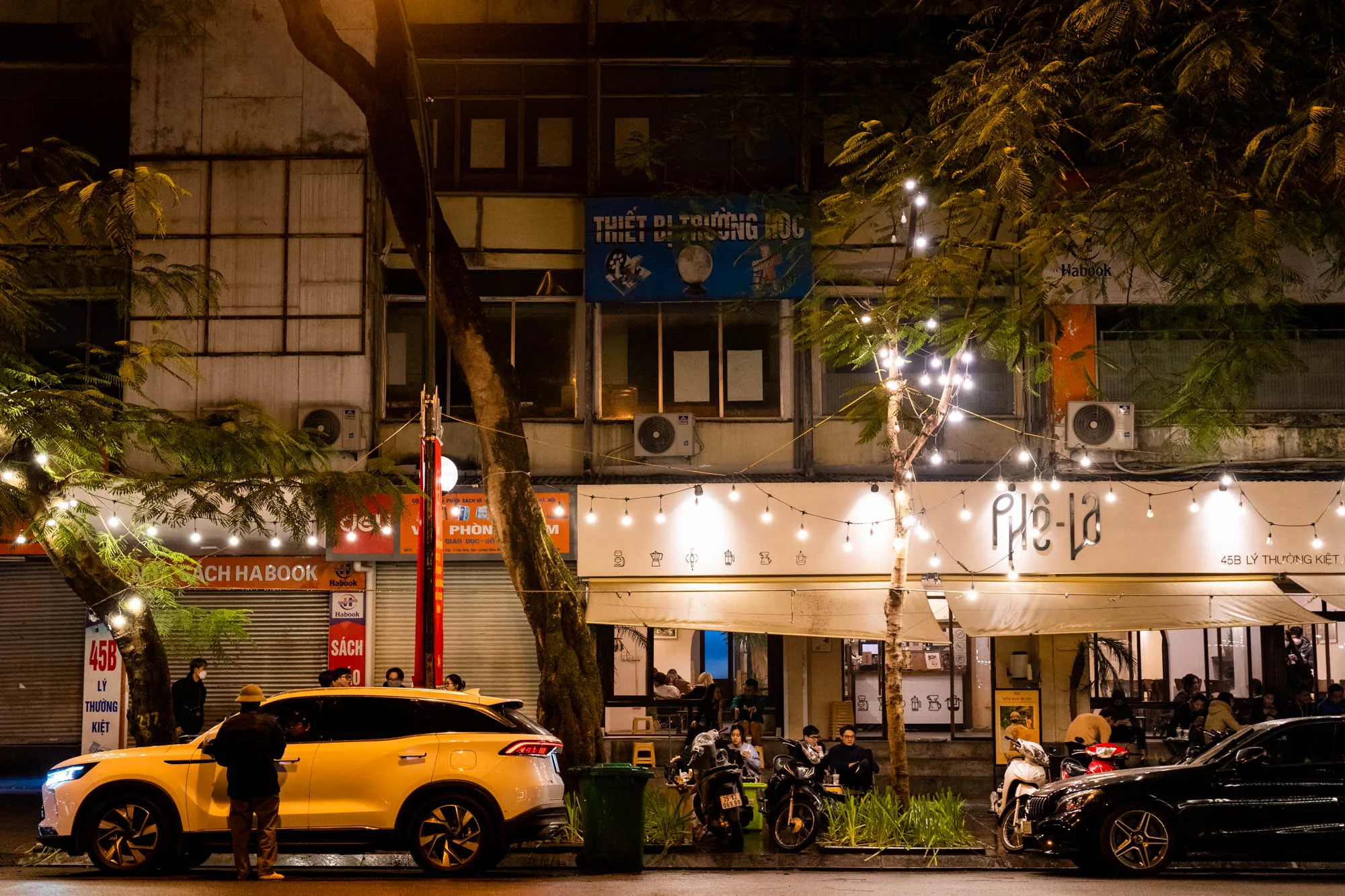 この写真はベトナムのハノイで撮影された夜の街並みを捉えています。画面中央には「Phê-la」という看板を掲げたカフェがあり、暖かい光を放つ電球のガーランドが飾られています。カフェの内部には人々が座って談笑している様子が見え、賑やかな雰囲気が伝わってきます。  写真の左側には「HA BOOK」と書かれた書店があり、その前にはクリーム色のSUVが停車しています。車に向かって話しかけている人物がいます。 道路にはスクーターや車が停まっており、ハノイの日常的な光景が写し出されています。街路樹がライトアップされており、夜の雰囲気をより一層引き立てています。建物の壁にはエアコンの室外機が設置されており、生活感を感じさせます。全体的に、ハノイの活気ある夜の様子が伝わってくる写真です。