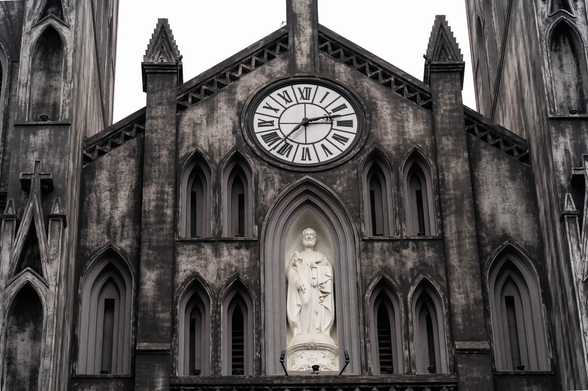 この写真はベトナムのハノイにある教会の一部を捉えたものです。教会はゴシック様式で、全体的に灰色がかった色調です。建物の表面は、年月を経たような、または意図的に古びたような質感を持っています。中央には大きな円形の時計があり、ローマ数字が使われています。その下には白い彫像が安置されています。彫像は人物像で、衣服を身につけています。建物の壁にはアーチ型の窓が整然と並んでいます。全体的に、歴史を感じさせる荘厳な雰囲気を醸し出しています。空は白く、建物の色調と相まって、落ち着いた印象を与えています。