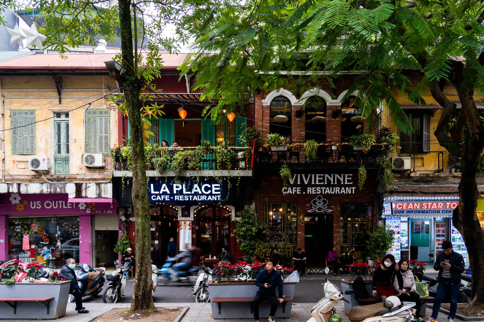 写真はベトナムのハノイで撮影された、活気のある通りを捉えています。画面中央には「LA PLACE」と「VIVIENNE」という名前のカフェ・レストランが隣接して建っています。それぞれの建物の外観は異なり、「LA PLACE」は濃い青色の看板が目を引く一方、「VIVIENNE」はレンガ造りで、クラシックな雰囲気を醸し出しています。両店のバルコニーには緑豊かな植物が飾られており、趣のある装飾となっています。道沿いにはベンチが設置され、人々が休憩したり談笑したりする姿が見られます。また、ベトナムらしい風景として、多くのバイクが停められており、生活感があふれています。左側には「US OUTLET」というショップがあり、ピンク色の外観が目を引きます。右側には旅行代理店の看板が見え、外国人観光客向けの情報を発信しているようです。全体的に、賑やかで活気があり、ハノイの日常の風景が垣間見える写真です。