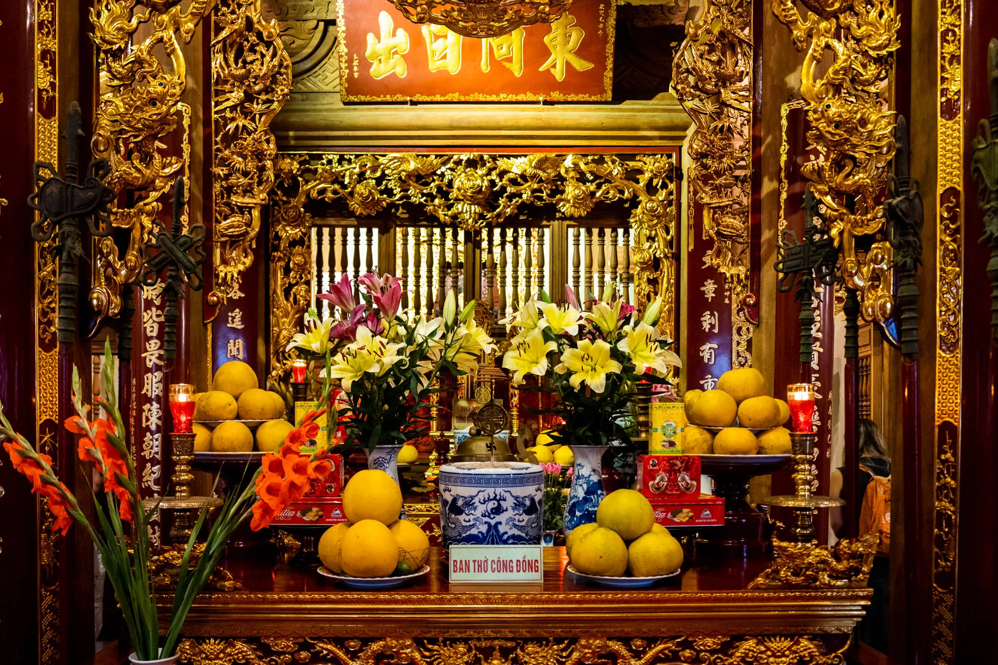 写真はベトナムのハノイで撮影された、豪華な祭壇の様子を捉えています。全体的に赤と金色を基調とした装飾が施されており、伝統的な雰囲気が漂っています。  祭壇の中央には、黄色の大きな果物（おそらく柑橘類）が複数皿に盛られており、その手前には青と白の陶器製の香炉が置かれています。香炉には陰陽のマークと龍の絵が描かれています。祭壇の上には、ピンクと黄色のユリの花束が左右対称に飾られています。  祭壇の後ろには、格子状の扉のようなものが見え、その上には金の装飾が施されています。扉の上には、漢字で書かれた看板が掲げられています（OCRの結果から「出國同東」と読めます）。  祭壇の左右には、赤色の柱があり、金色の龍などの装飾が施されています。柱には漢字のような文字が縦書きで書かれています（OCRの結果から「達聞前皆畏服陳朝紀章利有市」と読めます）。祭壇の左側には赤いグラジオラスの花が生けられています。左右には赤い蝋燭が灯されています。  写真全体から、この祭壇が重要な宗教的、文化的意義を持つ場所であることが伺えます。豪華な装飾や供え物から、信仰の深さや敬意が感じられます。
