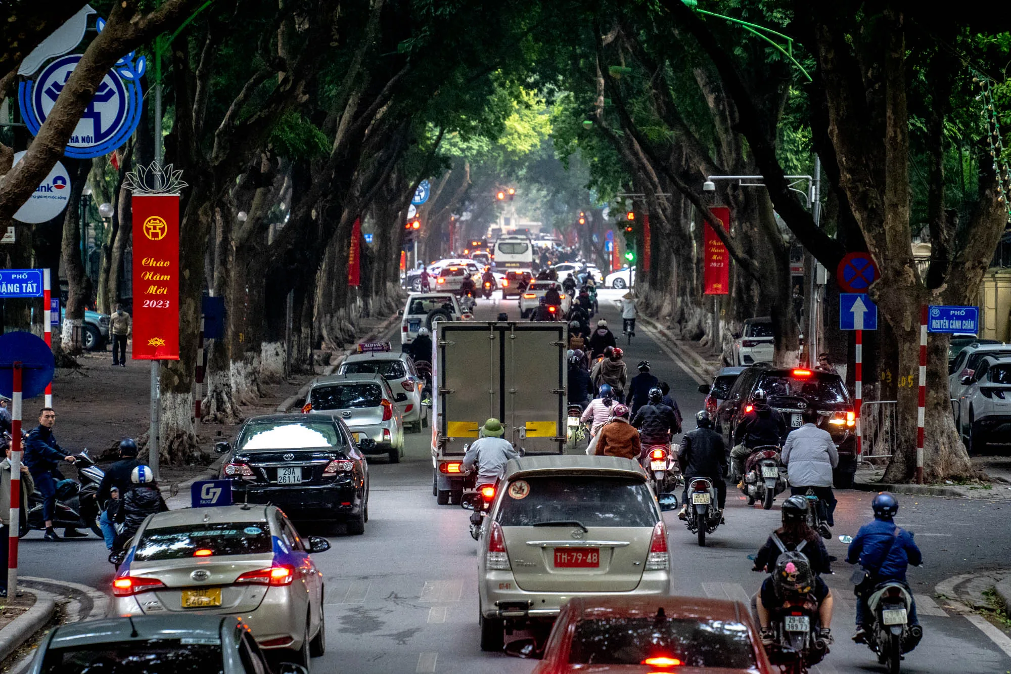 この写真はベトナムのハノイで撮影された、通りの風景です。画面奥へ続く道路は、両側に高く生い茂る緑豊かな街路樹によって覆われ、トンネルのような景観を作り出しています。  道路には、自動車やバイクがひしめき合い、交通量が多い様子が伺えます。バイクに乗る人々は様々な服装をしており、日常の風景を切り取ったような自然な様子です。車のナンバープレートも写っており、ハノイの街並みであることがわかります。道路脇にはベトナム語で書かれた看板や標識が立っており、その地域特有の文化や雰囲気を醸し出しています。赤い垂れ幕には「Chào Năm Mới 2023」(新年おめでとう2023)と書かれており、撮影時期が2023年の新年であることを示唆しています。全体的に、ハノイの活気ある日常を描写した写真と言えるでしょう。