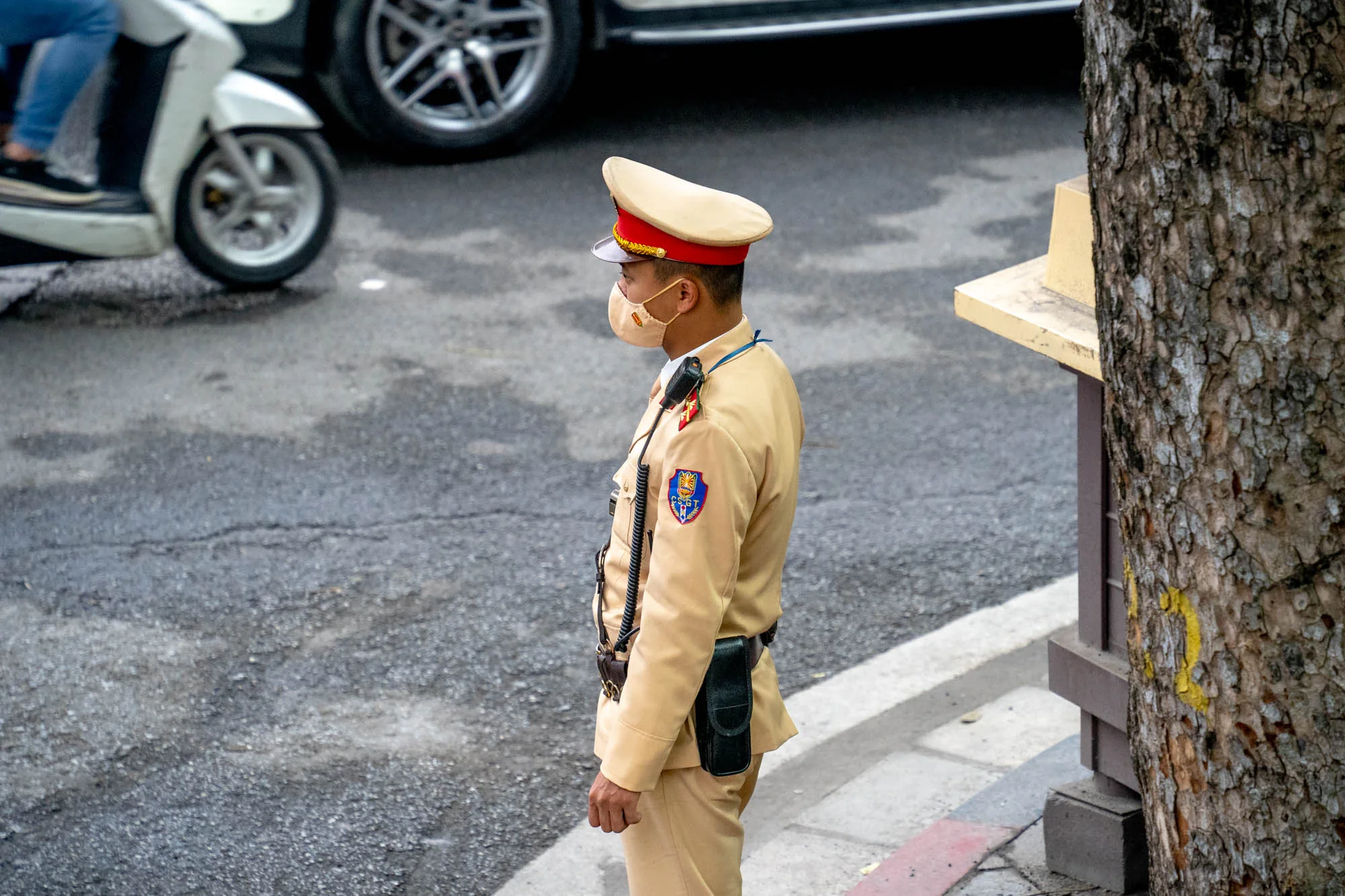 ベトナムのハノイで撮影された写真です。画面中央には、制服を着た男性の警官が写っています。警官は、ベージュ色の制服に赤いラインの入った帽子を着用し、マスクをしています。肩には無線機のようなものがかけられ、腰にはホルスターが装着されています。警官は横を向いており、道路を見ているようです。背景には、アスファルトの道路、白いスクーターの一部、車のタイヤ、そして木の幹が見えます。道路は少し濡れているように見え、全体的に落ち着いた雰囲気の写真です。警官の制服の左腕には、紋章のようなものが縫い付けられています。
