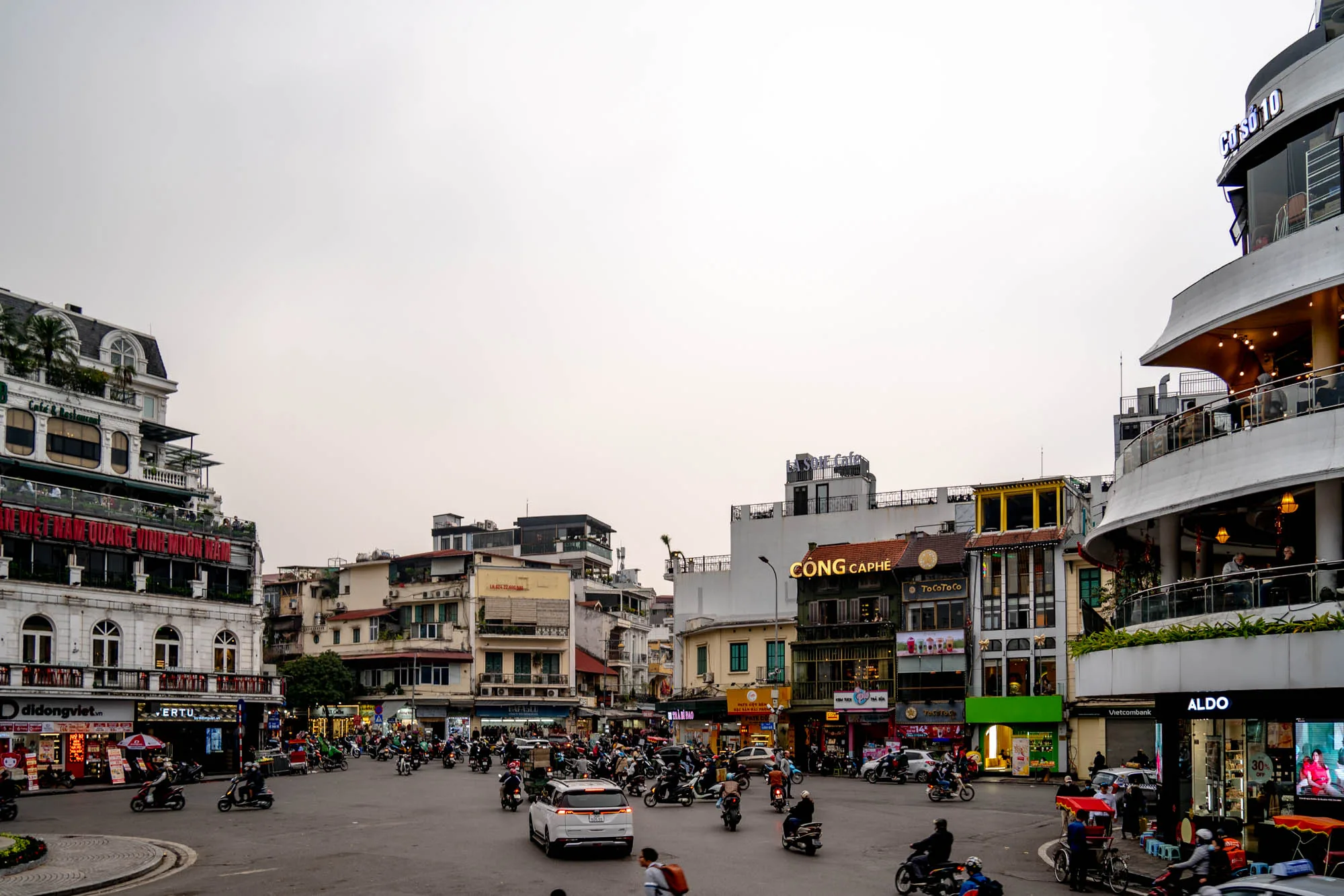 写真はベトナムのハノイで撮影されたもので、交差点の様子が写っています。空はどんよりと曇っており、時間帯は夕暮れ時かと思われます。交差点には、無数のバイクや車が入り乱れ、活気に満ちた混沌とした雰囲気が伝わってきます。周囲には様々な高さの建物が立ち並び、それぞれに異なるデザインや看板が掲げられています。例えば、「CONG CAPHE」や「ALDO」といった店舗が見られます。建物の上部にはカフェやレストランがあり、人々が食事を楽しんでいる様子が伺えます。通りの歩道には、広告や店舗が並び、非常に賑やかです。写真全体から、ハノイの街のエネルギーと多様性を感じることができます。