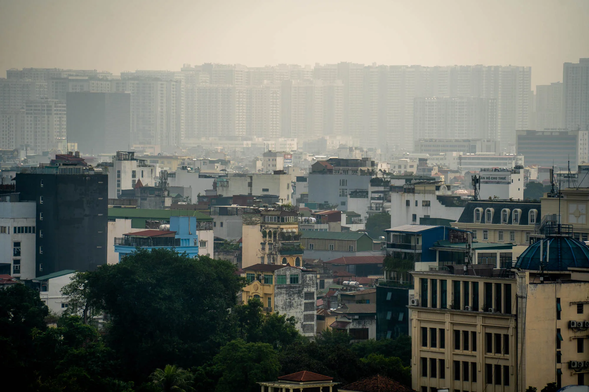 この写真はベトナムのハノイで撮影された都市の風景を捉えています。前景には緑豊かな木々が茂り、その奥には様々な高さと色の建物が密集して建ち並んでいます。建物は、古いものから新しいものまで、様々な様式が見られます。一部の建物はコロニアル様式のような、特徴的な屋根や窓を持つものもあります。また、新しい高層ビルも混ざっており、都市の発展と変化を感じさせます。  写真全体には、大気汚染か霧のためか、靄がかかっており、遠くの建物はぼやけて見えます。この靄が、写真に独特の雰囲気を与え、都市の喧騒を和らげているようにも見えます。空は白く、全体的に落ち着いた色調の写真です。都市の密度と多様性が感じられる一枚と言えるでしょう。