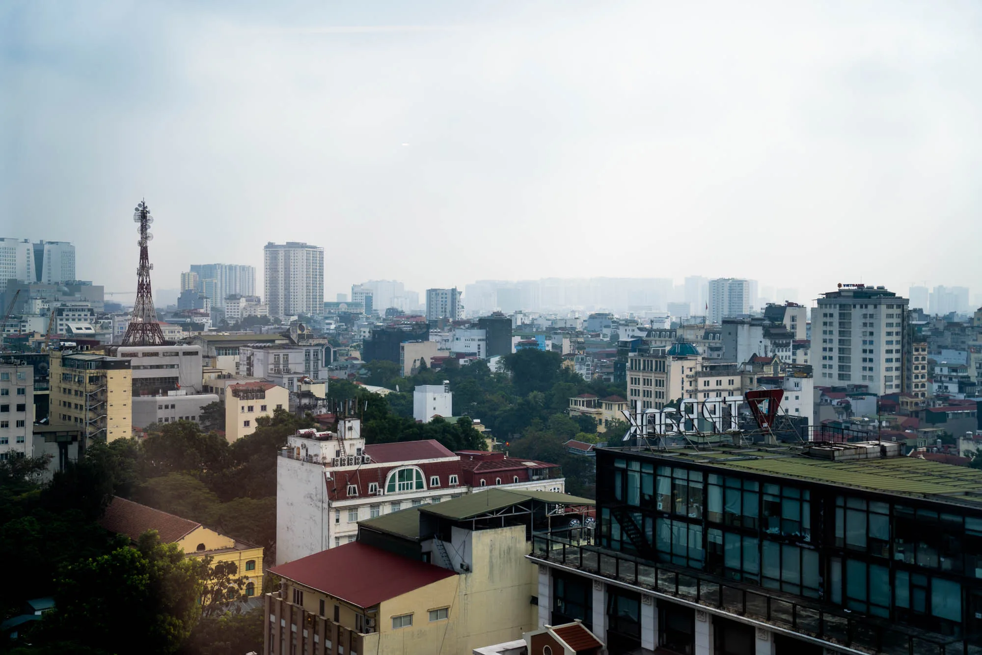 写真は、ベトナムのハノイで撮影された都市の風景です。やや霞がかった空の下、様々な高さの建物が密集して建ち並んでいる様子が捉えられています。画面手前には、屋上緑化されたモダンなビルが一部写っており、その向こうには赤茶色の屋根や緑豊かな木々が見えます。中央奥には、ハノイのランドマークである鉄塔がそびえ立っています。さらに遠くには、高層ビル群が霞の中に浮かび上がり、奥行きのある景観を形成しています。全体的に、都市の活気とベトナムの独特な雰囲気が伝わる写真です。